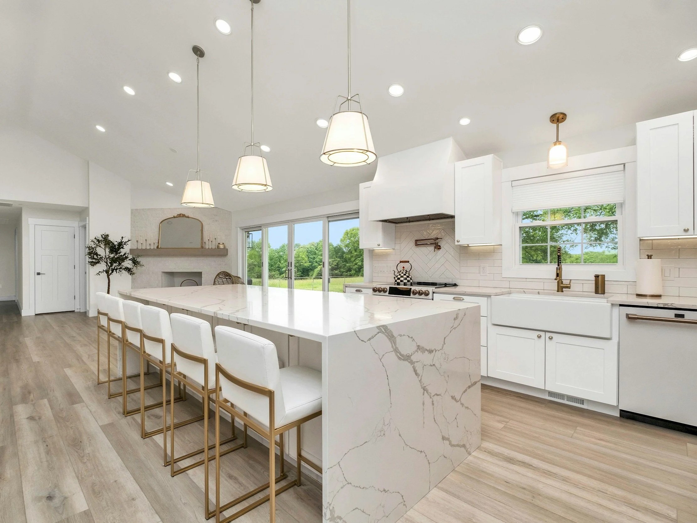 Modern white kitchen with an island, gold chairs, and large windows showing green outdoor scenery.