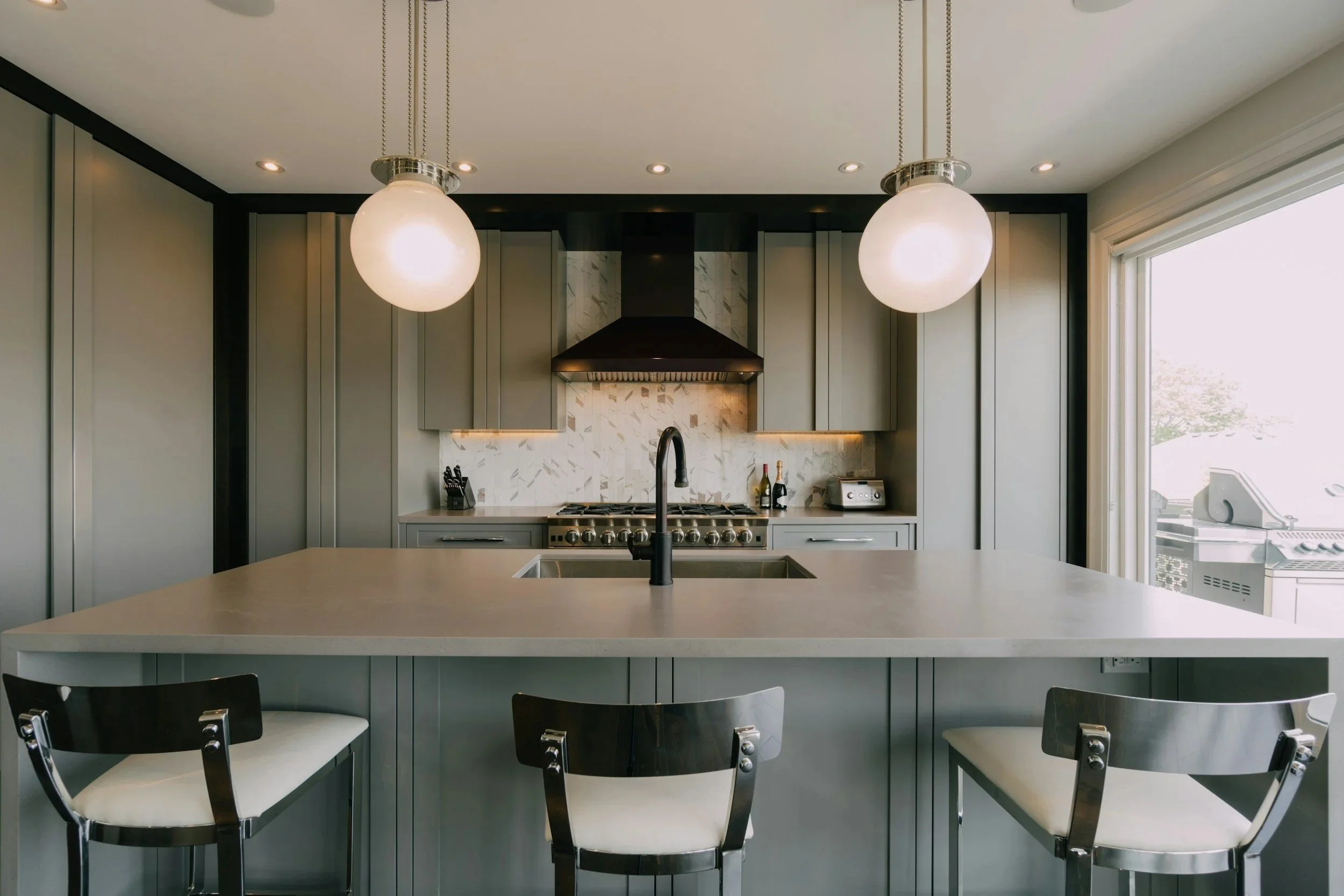 Modern kitchen with gray cabinets, two large pendant lights, marble backsplash, black faucet, stainless steel appliances, and a large kitchen island with seating.