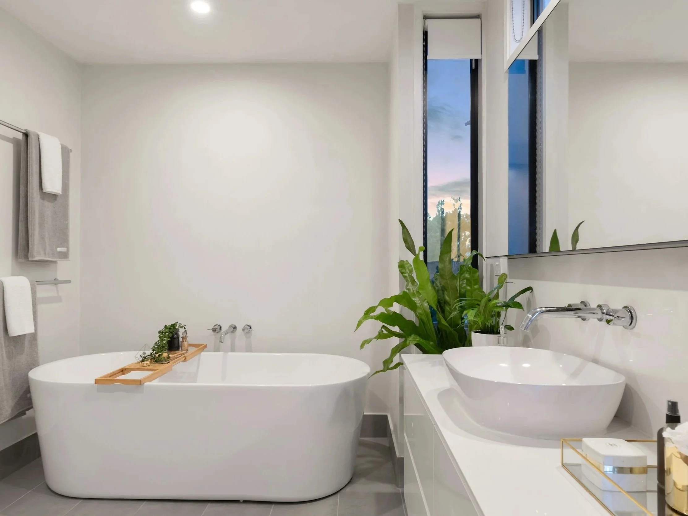 Modern bathroom with a white bathtub, a large mirror, a vessel sink, green plants, and white towels on a towel rack, with large windows showing a sunset sky.