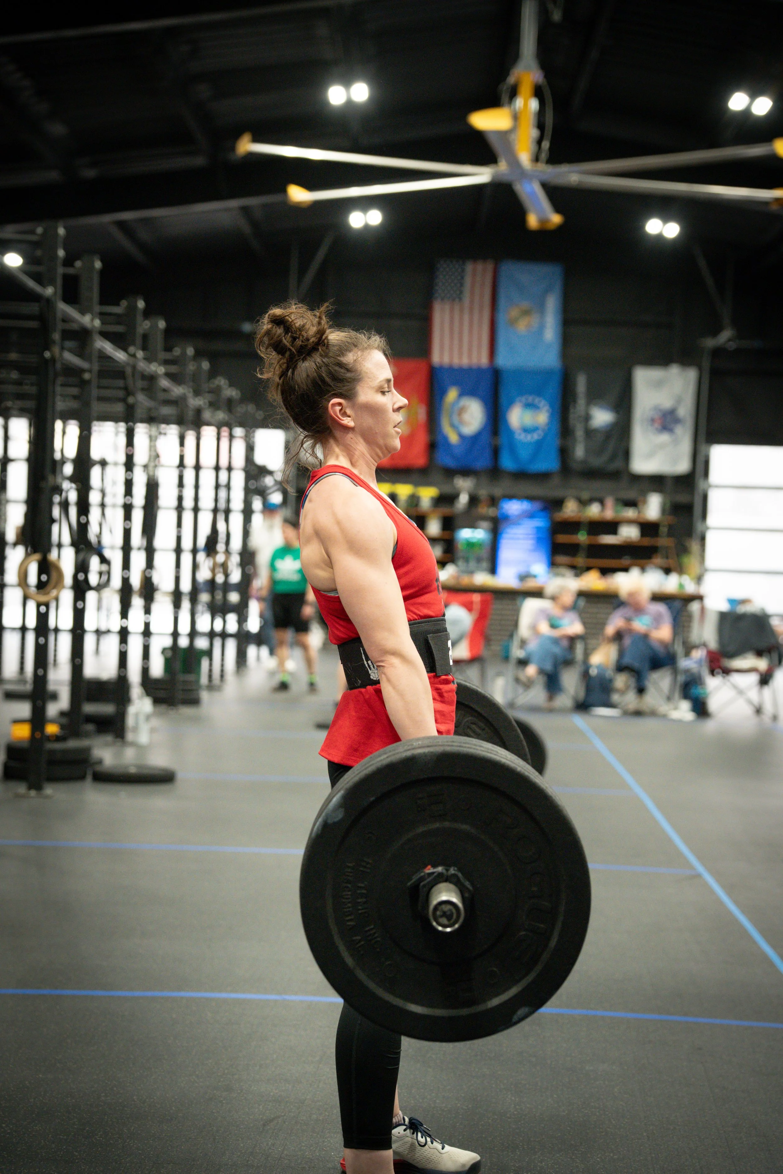 A woman in a red workout outfit lifting a barbell in a gym with flags hanging on the wall.