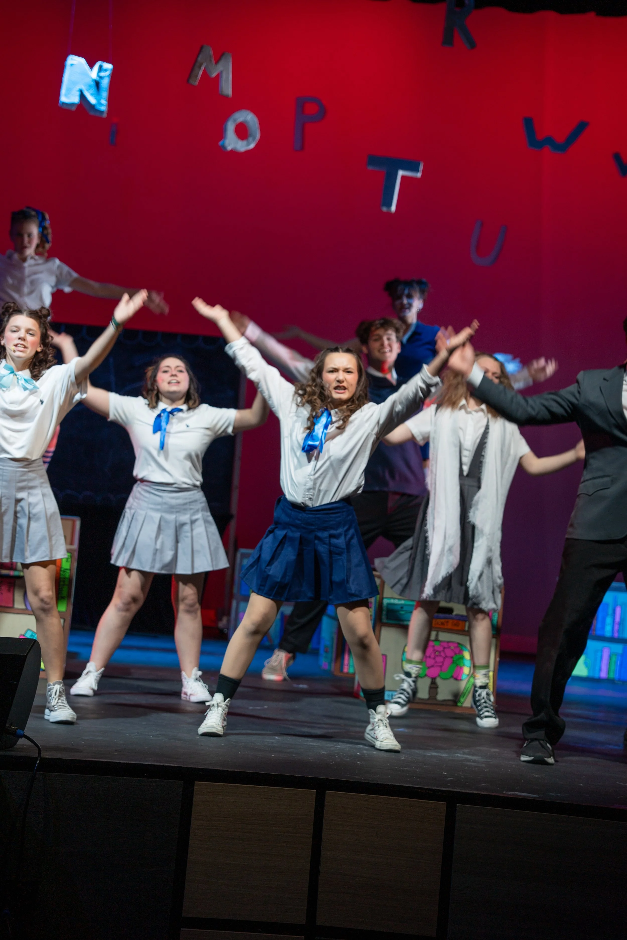 Young girls and a woman performing on stage in school uniforms with blue ties, some with painted faces, under colorful lights, with a red backdrop and alphabet letters hanging overhead.
