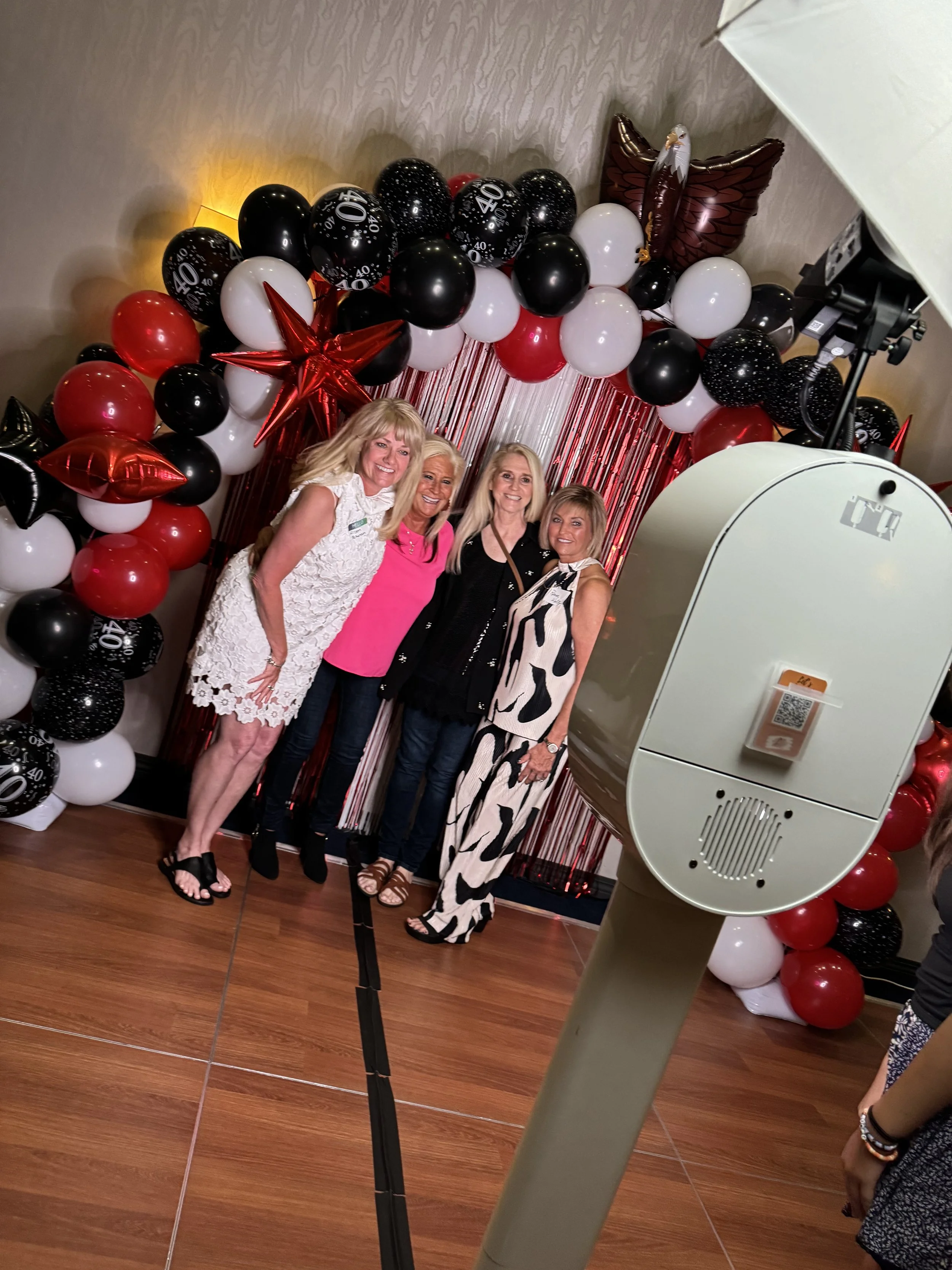 Four women smiling and posing in front of a balloon backdrop with red, black, and white balloons, and a shiny red fringe curtain. A photo booth camera is in the foreground.