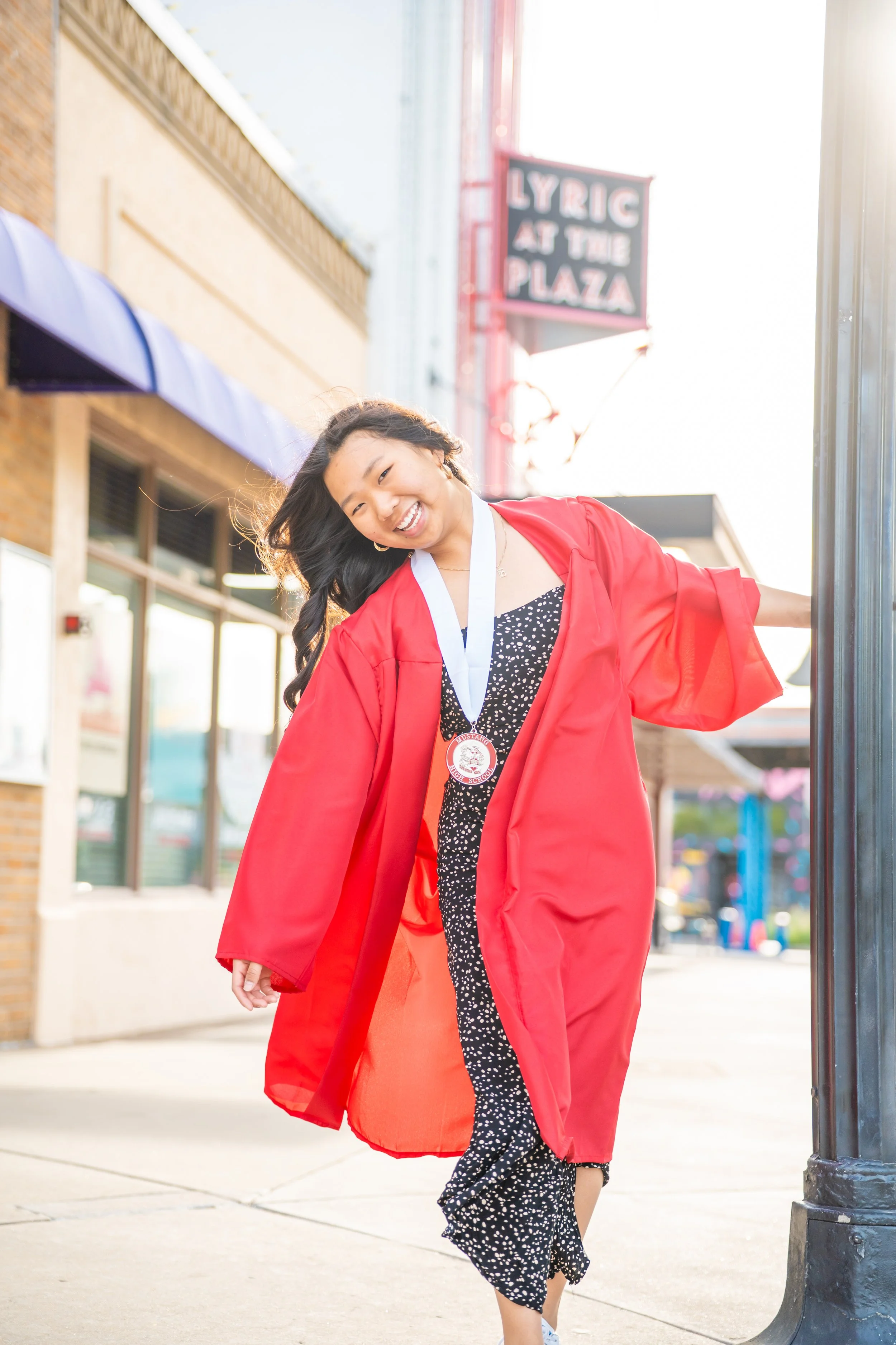 Young woman in a red graduation gown smiling and walking outdoors on a bright day, wearing a medal around her neck.
