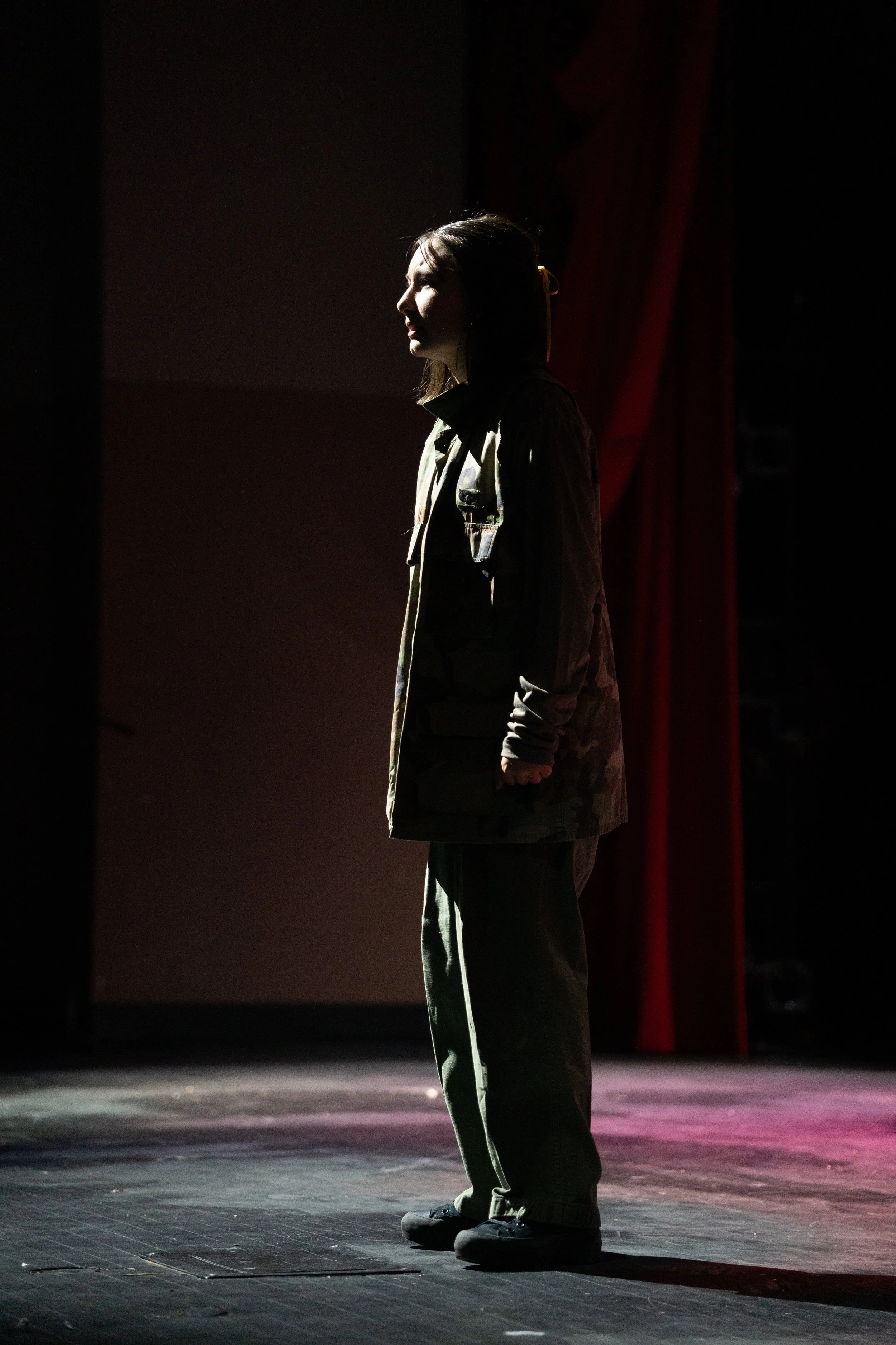 A woman standing on stage in profile, illuminated by a spotlight, with red curtains in the background.