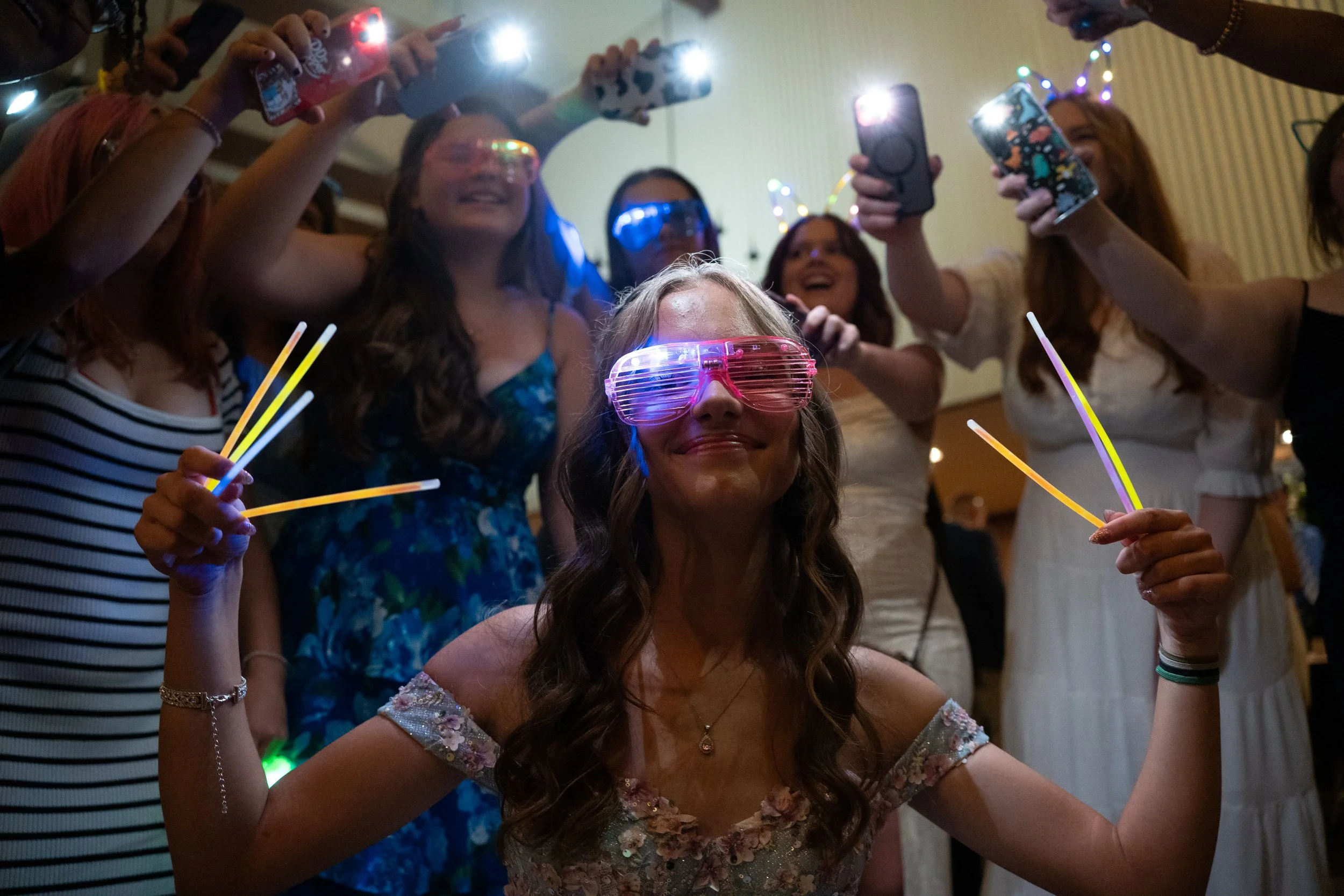 Woman with glow sticks and pink shutter shades smiling in the center, surrounded by friends taking photos and wearing glow accessories at a party.
