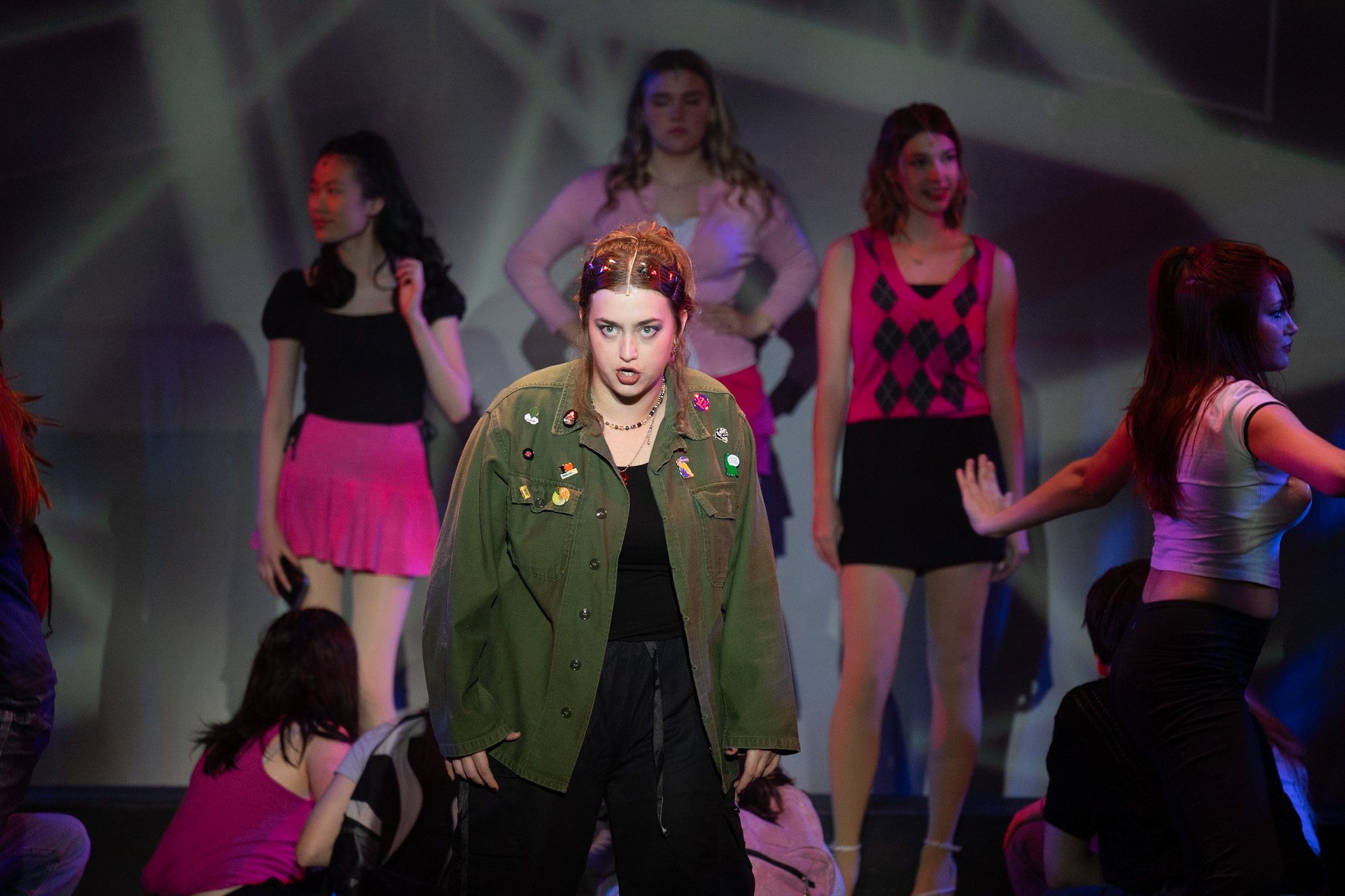 Young woman in a green jacket with pins, standing center stage, with other young women around her, some standing and some kneeling, under stage lighting.