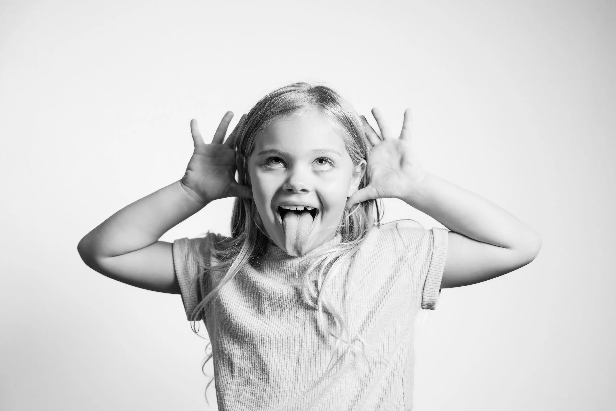 A young girl with long hair making a playful face, sticking out her tongue, with her hands cupped around her ears.