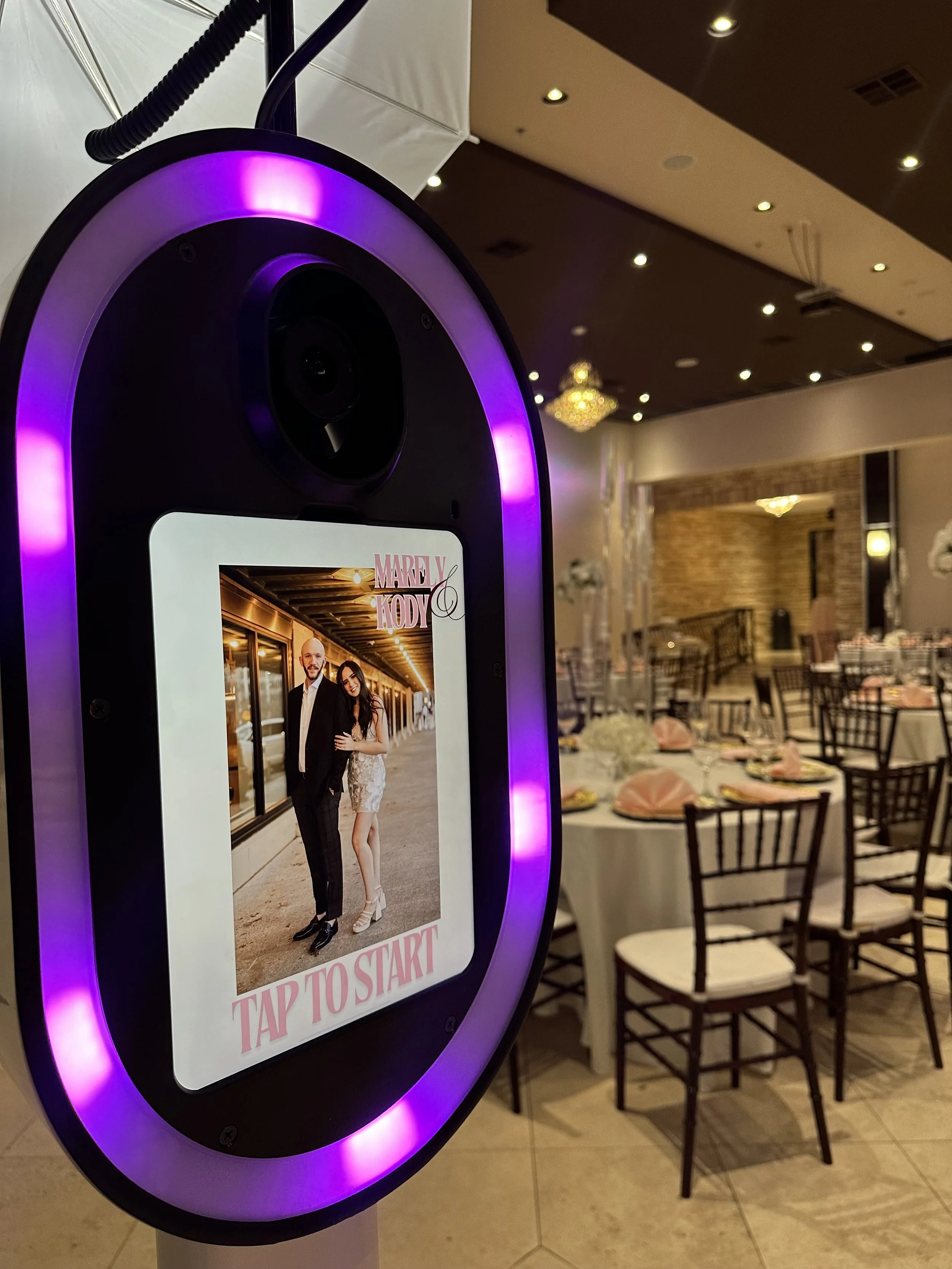 A photo booth screen showing a picture of a man and woman dressed in formal wear, with a decoratively set banquet hall in the background.