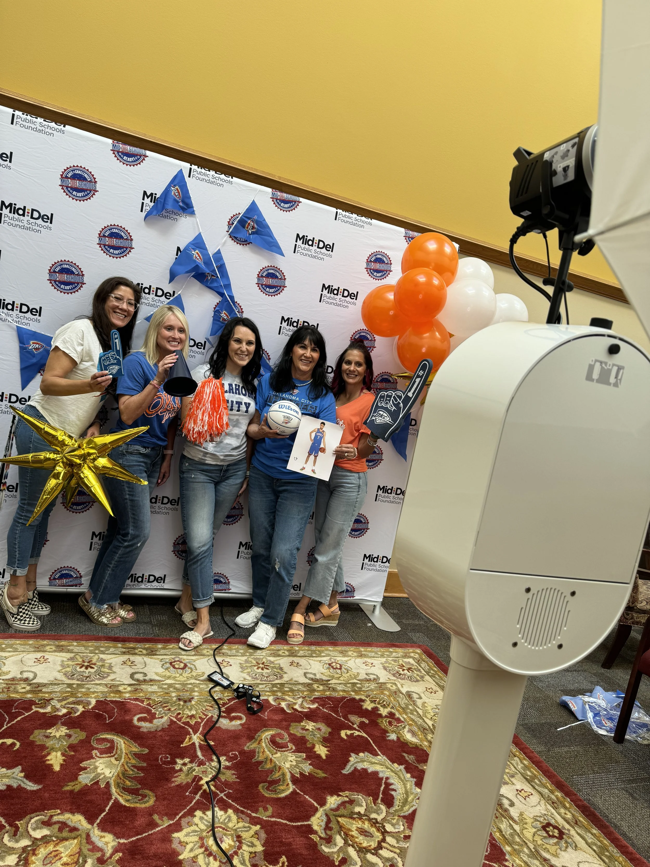 Group of women posing for a photo during a celebration event, with balloons, flags, and a backdrop with MidDel Public Schools Foundation logos.