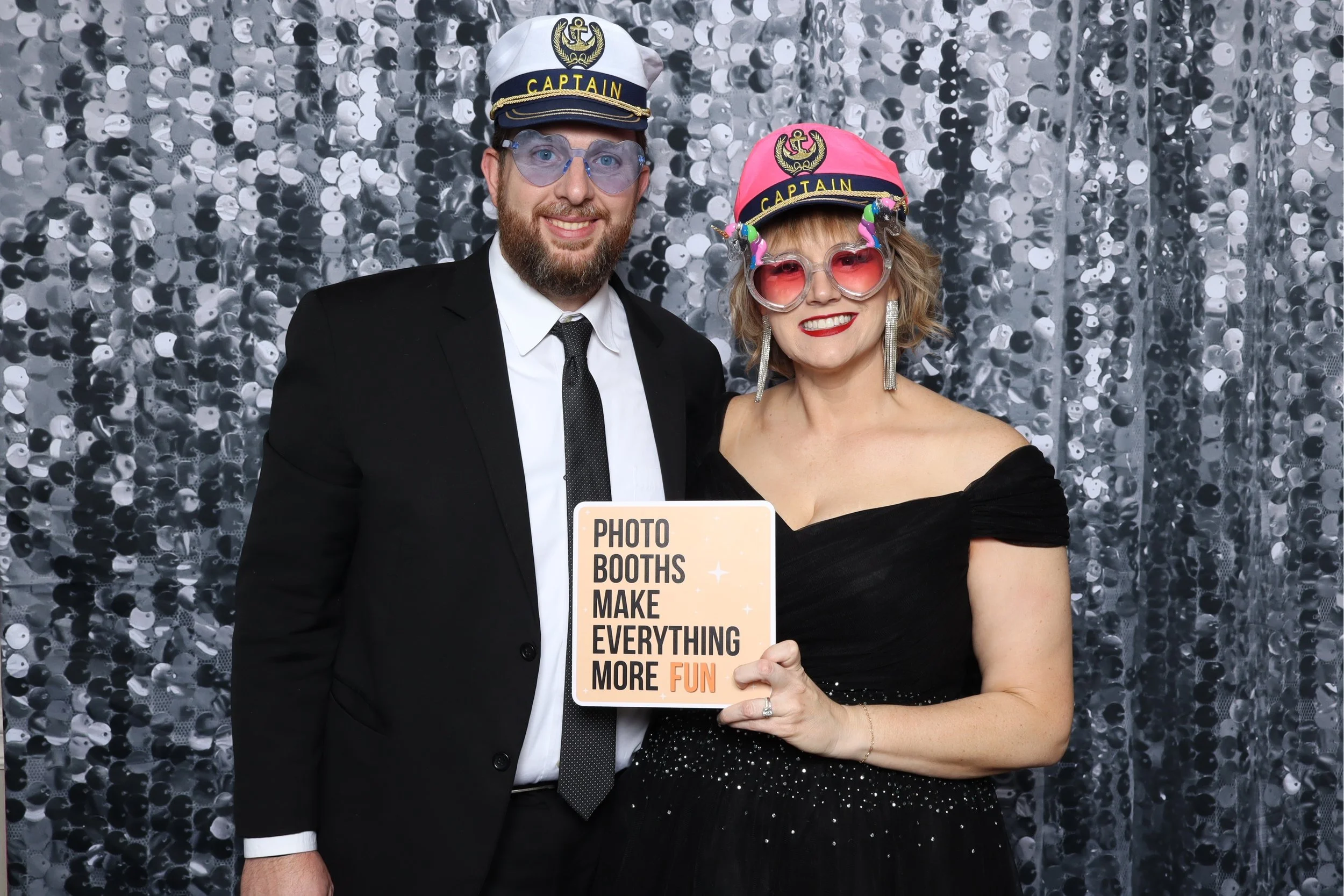 A man and woman at a photo booth, both wearing captain hats and fun sunglasses. The woman is holding a sign that reads, 'Photo booths make everything more fun.' They are dressed formally, with the man in a suit and tie, and the woman in an off-should