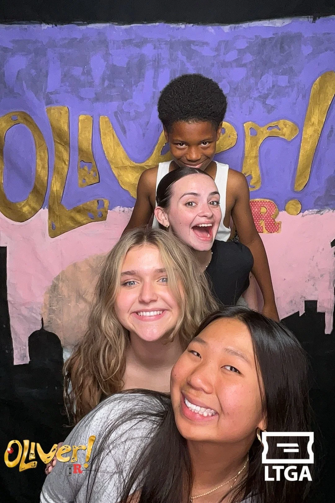 Four women and a young boy posing in front of a colorful backdrop with large gold and purple letters. The women are smiling and stacked in a pyramid style, with the boy at the top. The backdrop says 'Oliver!'