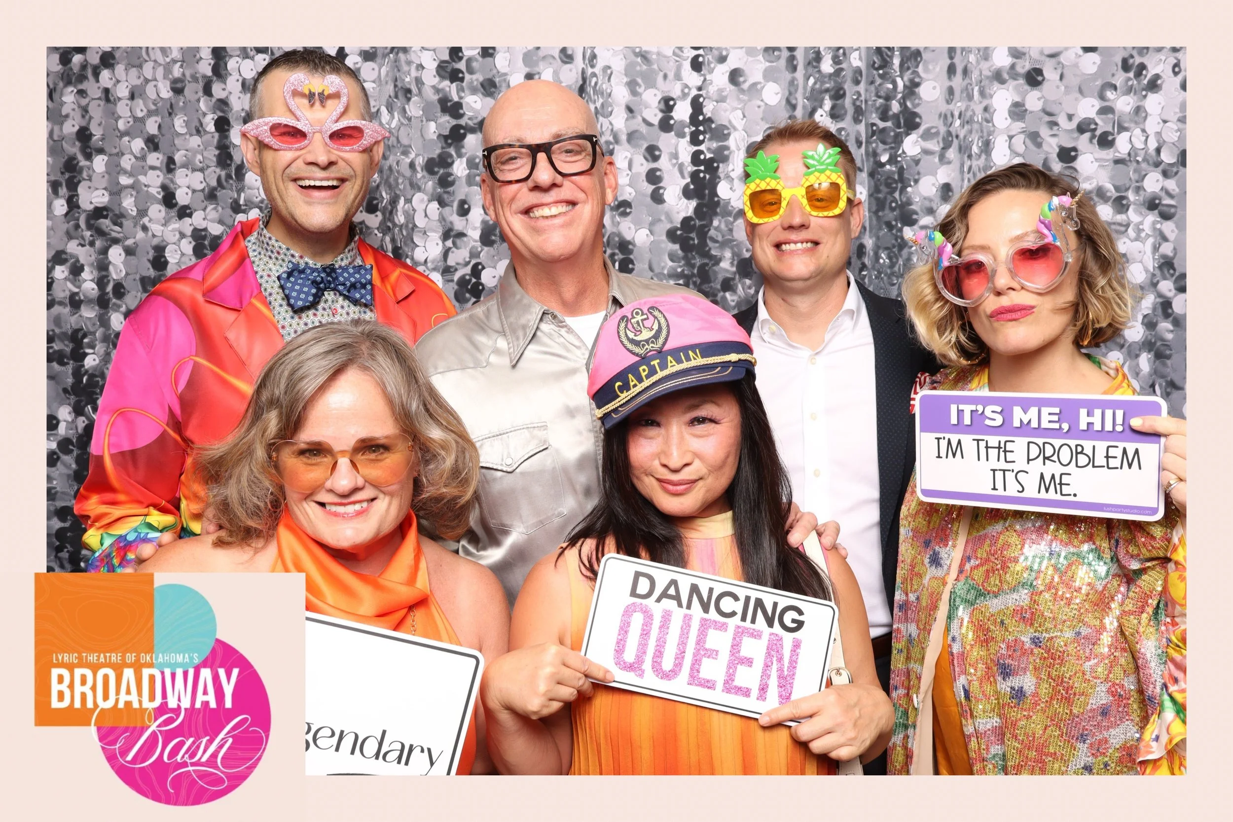 Group of six people at a Broadway-themed event, wearing colorful costumes and fun accessories, posing in front of a silver sequin backdrop, holding signs with playful messages.