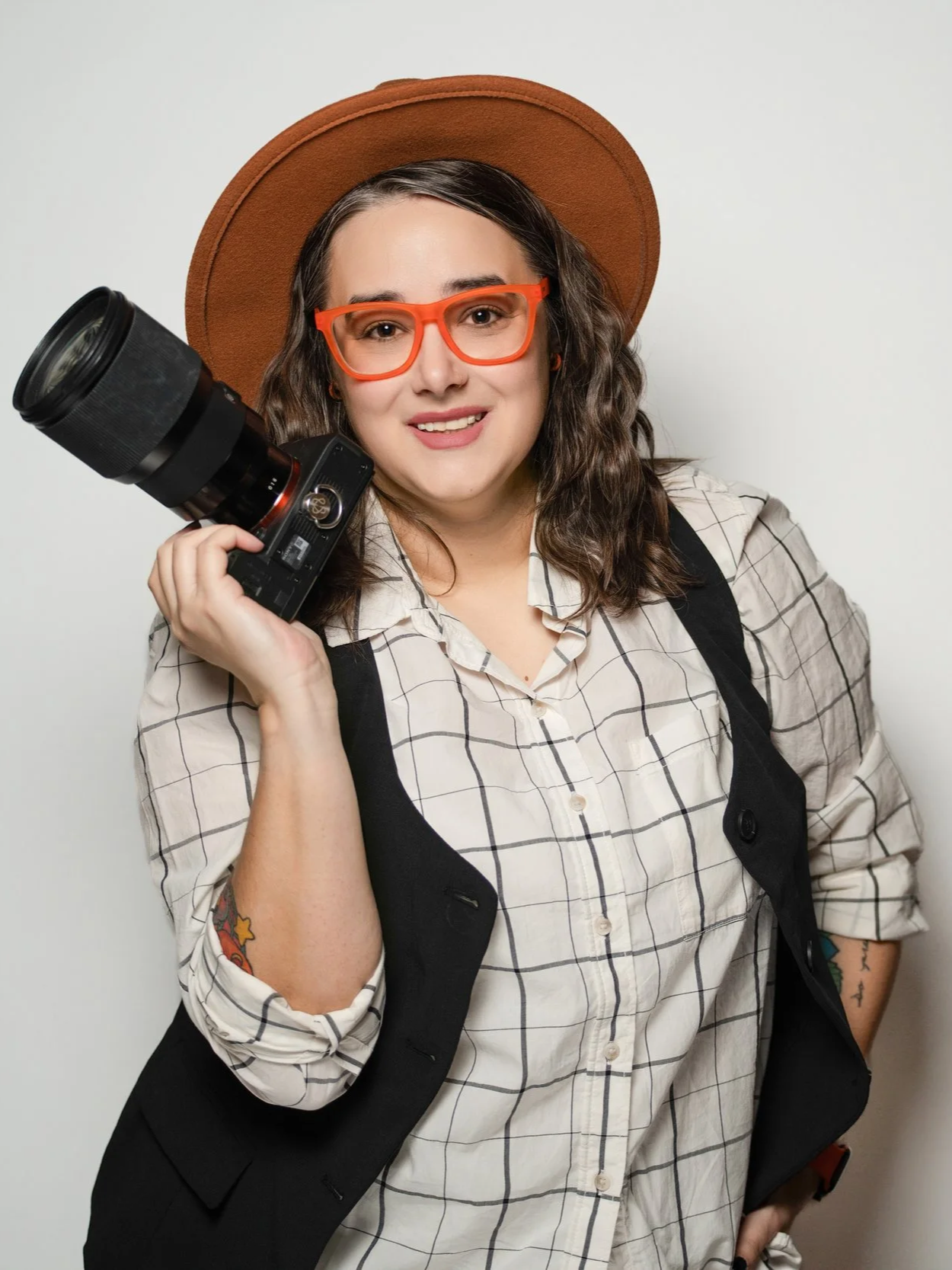 A woman with wavy dark hair wearing orange glasses, a tan hat, a beige plaid shirt, and a black vest, holding a professional camera with a large lens.