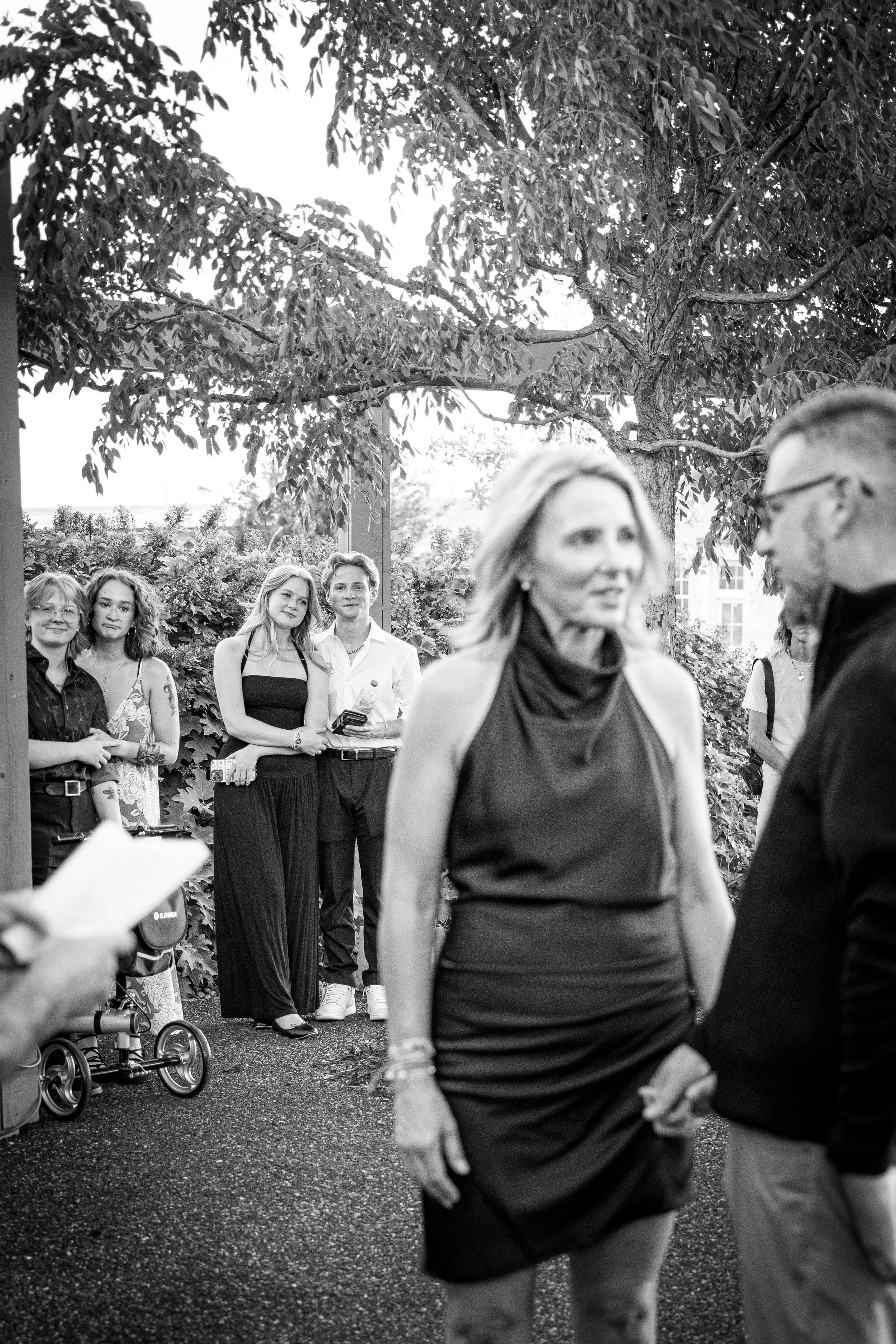 A black and white photo of a group of people outdoors, with a woman in a sleeveless dress and a man in glasses in the foreground, holding hands, while others observe from behind under a large tree.