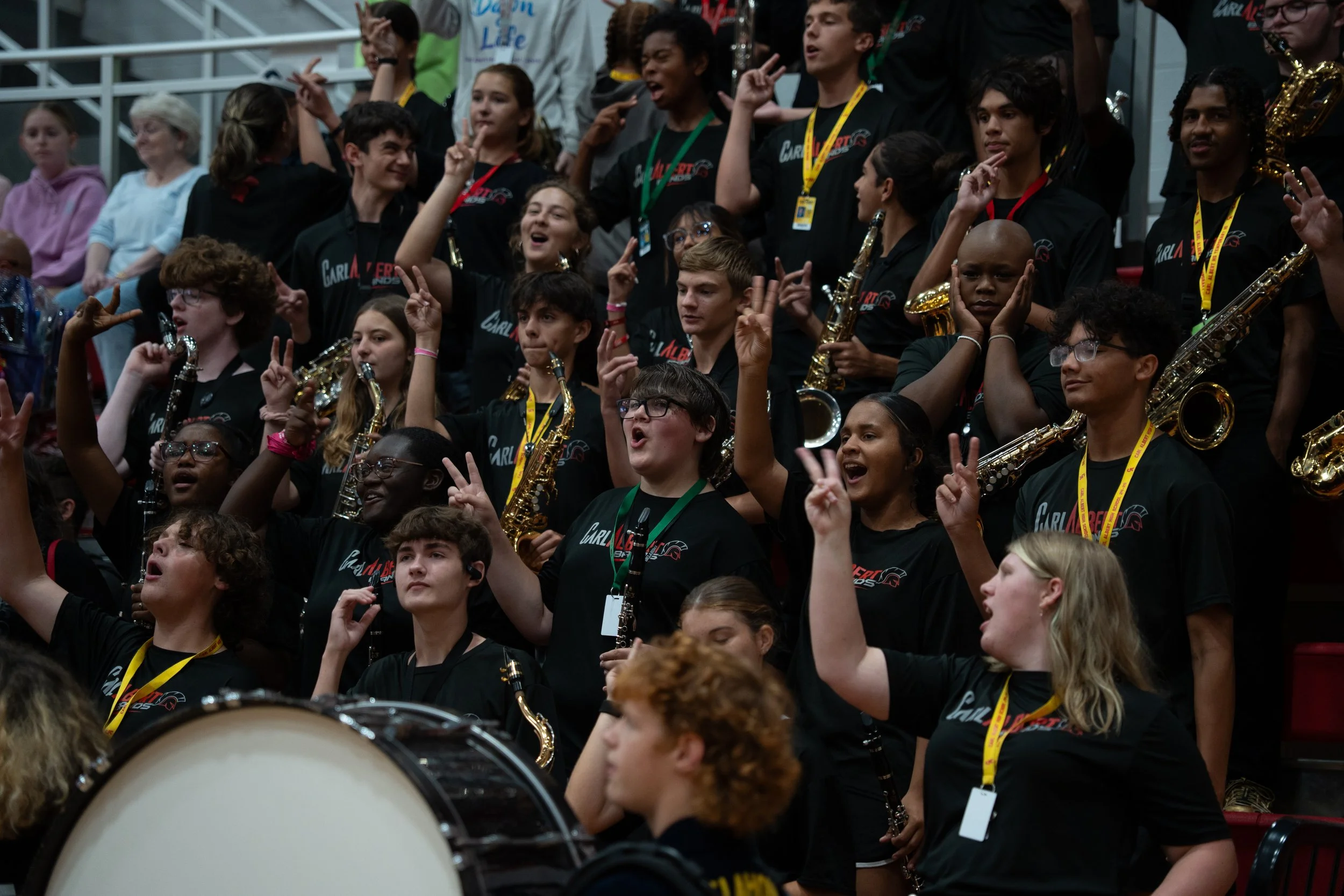 A large group of young musicians holding saxophones, participating in a band performance, wearing black shirts, some with yellow lanyards, in an indoor stadium or arena.