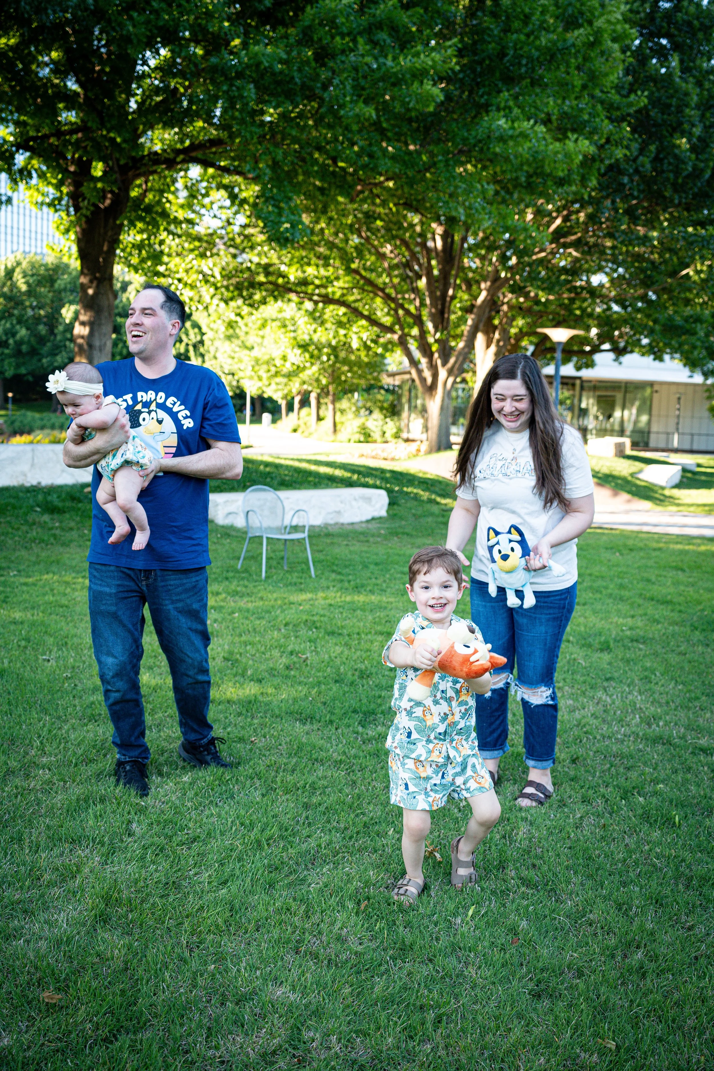 “Family walking together across a grassy park while a young child runs ahead smiling, holding a stuffed toy during a candid moment.”