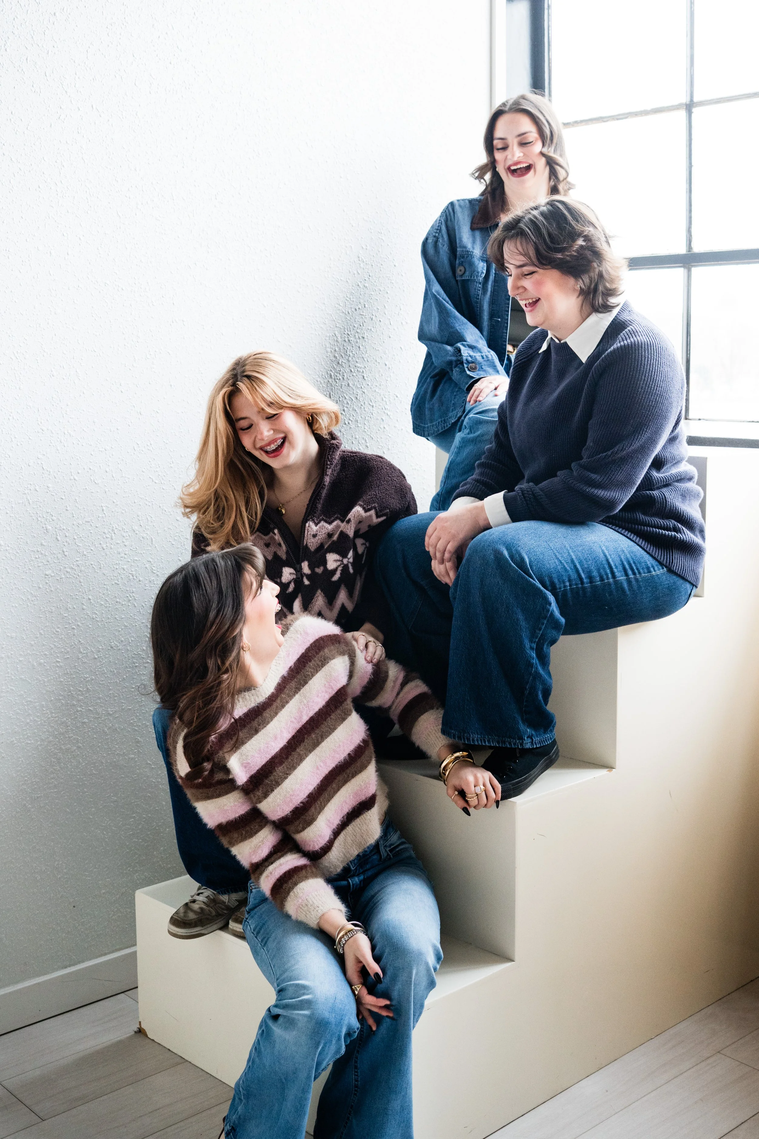 Four sisters laughing together while sitting on white tiered steps near a large window, captured in a natural light, candid moment