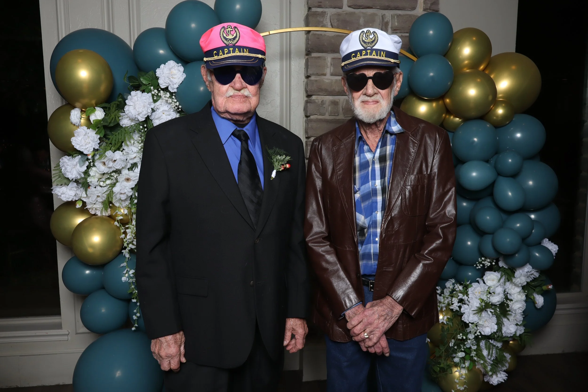 Two elderly men standing side by side in front of a balloon and floral decoration. Both are wearing captain hats with the word 'Captain' on them and dark sunglasses. The man on the left is dressed in a black suit with a blue shirt, black tie, and whi