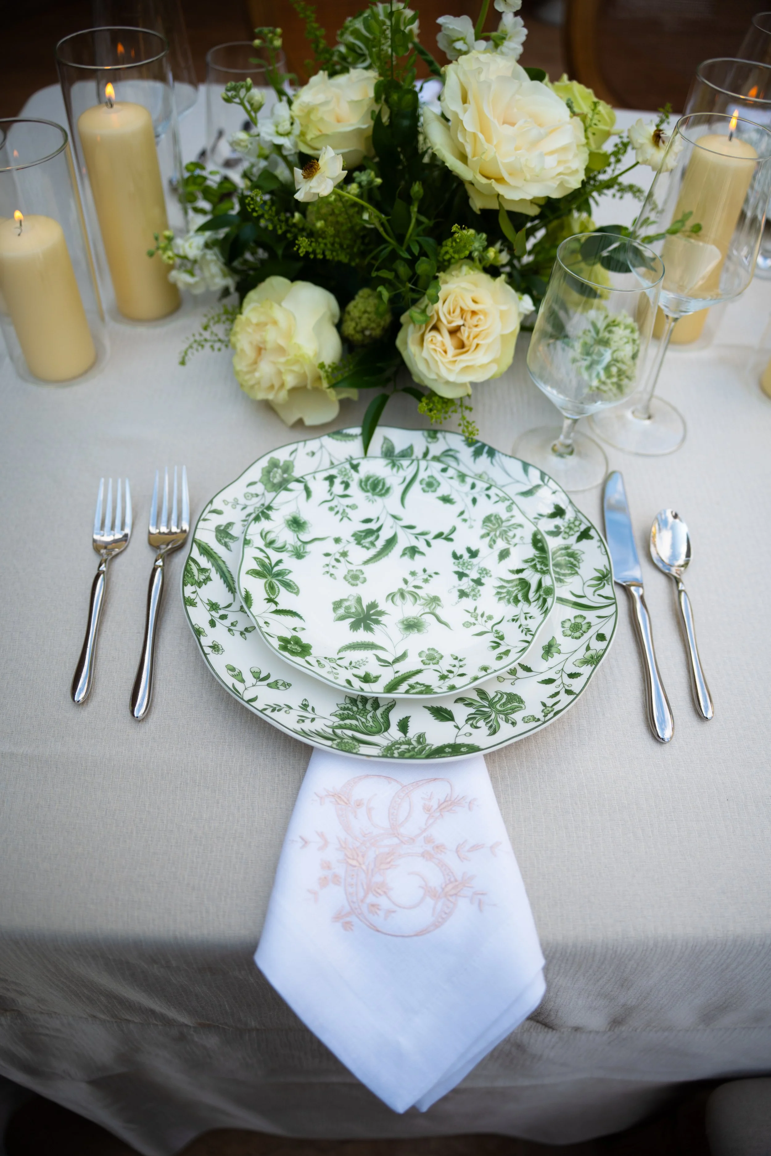 A formal dining table setting with a green and white floral patterned plate, a white embroidered napkin, silverware, empty wine glasses, candles, and a centerpiece with white and green flowers.