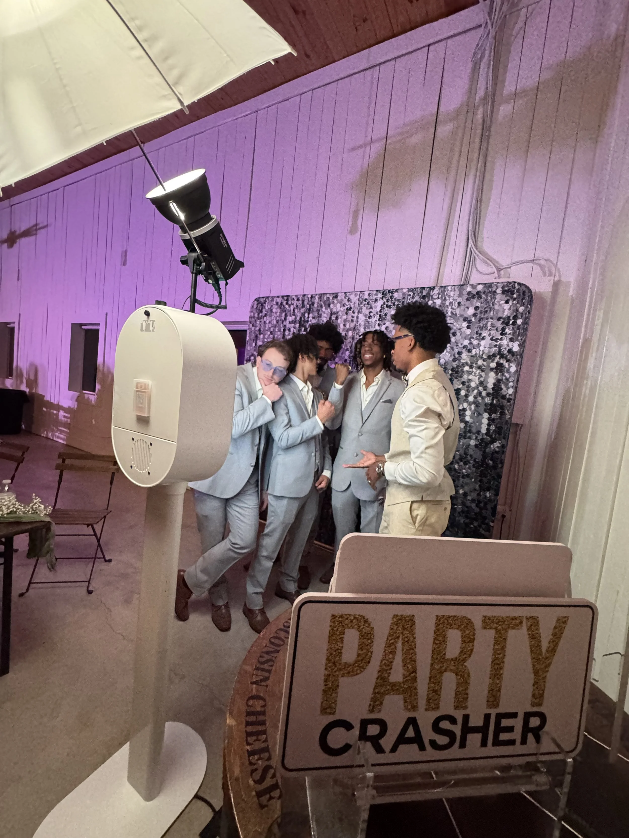 A group of five young adults dressed in light gray and cream suits taking selfies in front of a decorative backdrop at a party event. There is a large sign in the foreground that reads 'PARTY CRASHER' and a ring light setup.