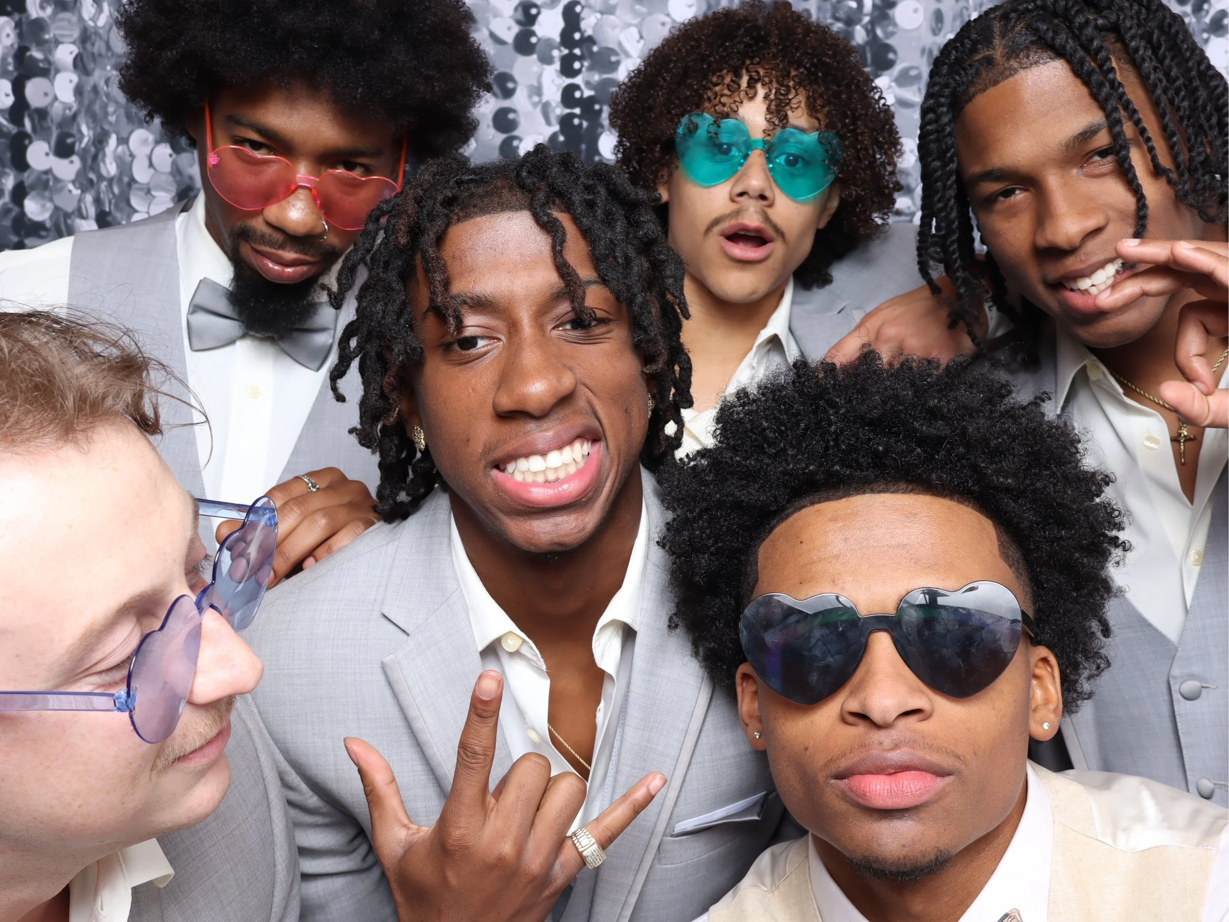 Group of seven people dressed in stylish suits with some wearing sunglasses and making expressive gestures, posing for a photo in front of a decorative backdrop.