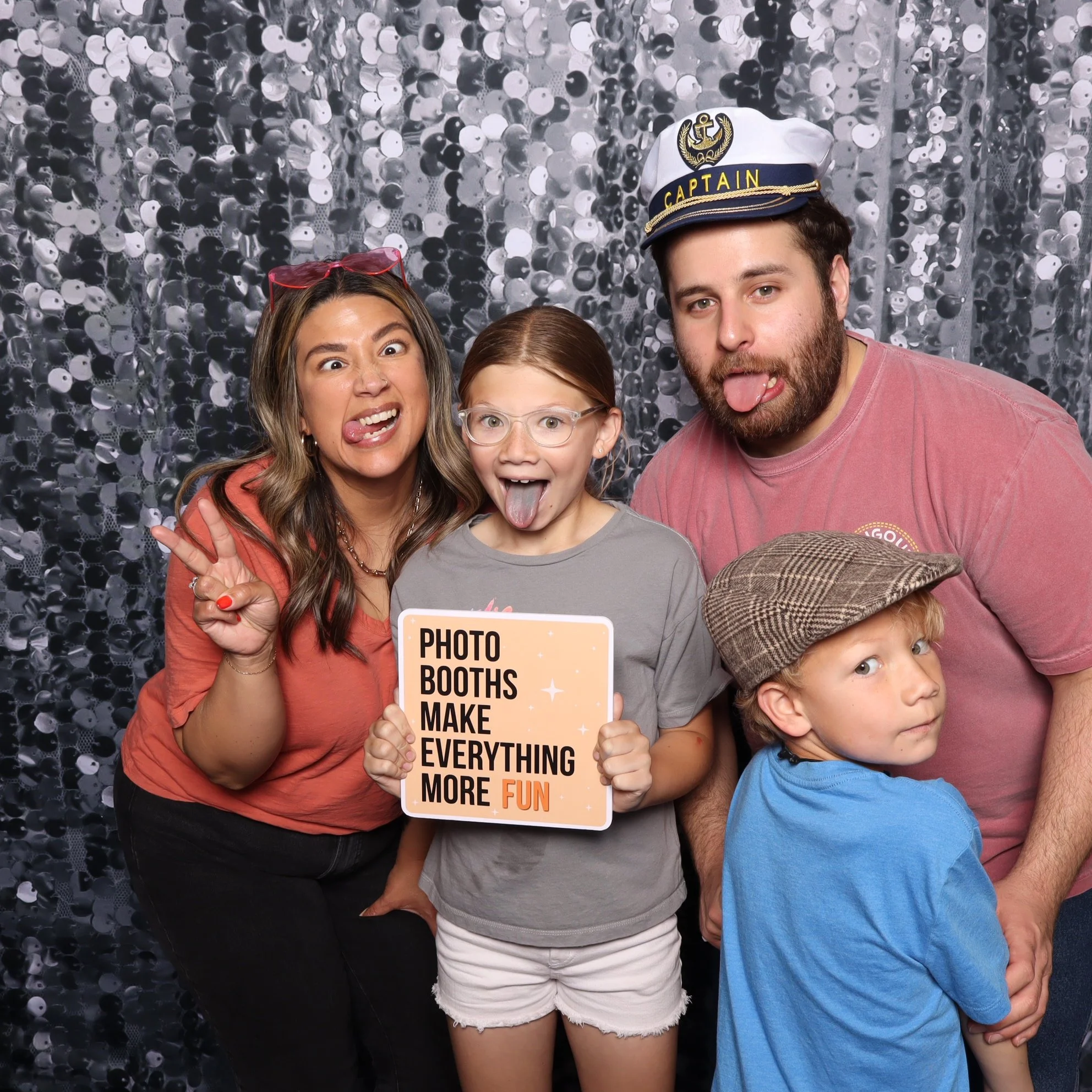 Group of four people at a photo booth, making silly faces and holding a sign that says "Photo booths make everything more fun", with a sparkly sequin backdrop.