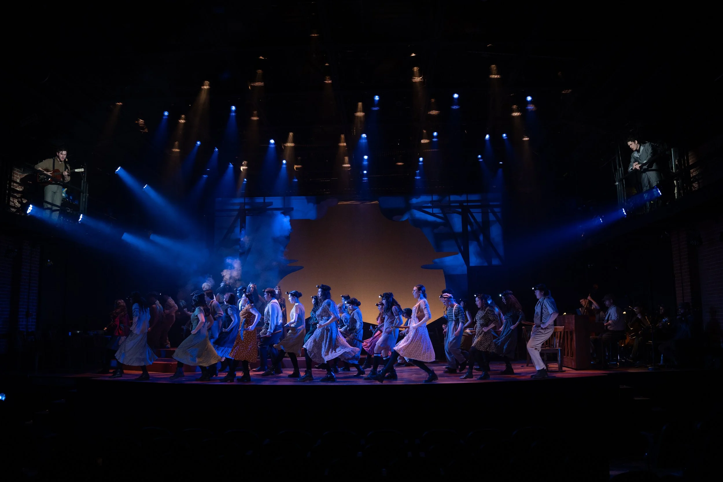 Stage scene with performers in vintage costumes dancing, with musicians and guitarist on either side, stage lit with blue and yellow lights, theatrical set backdrop.