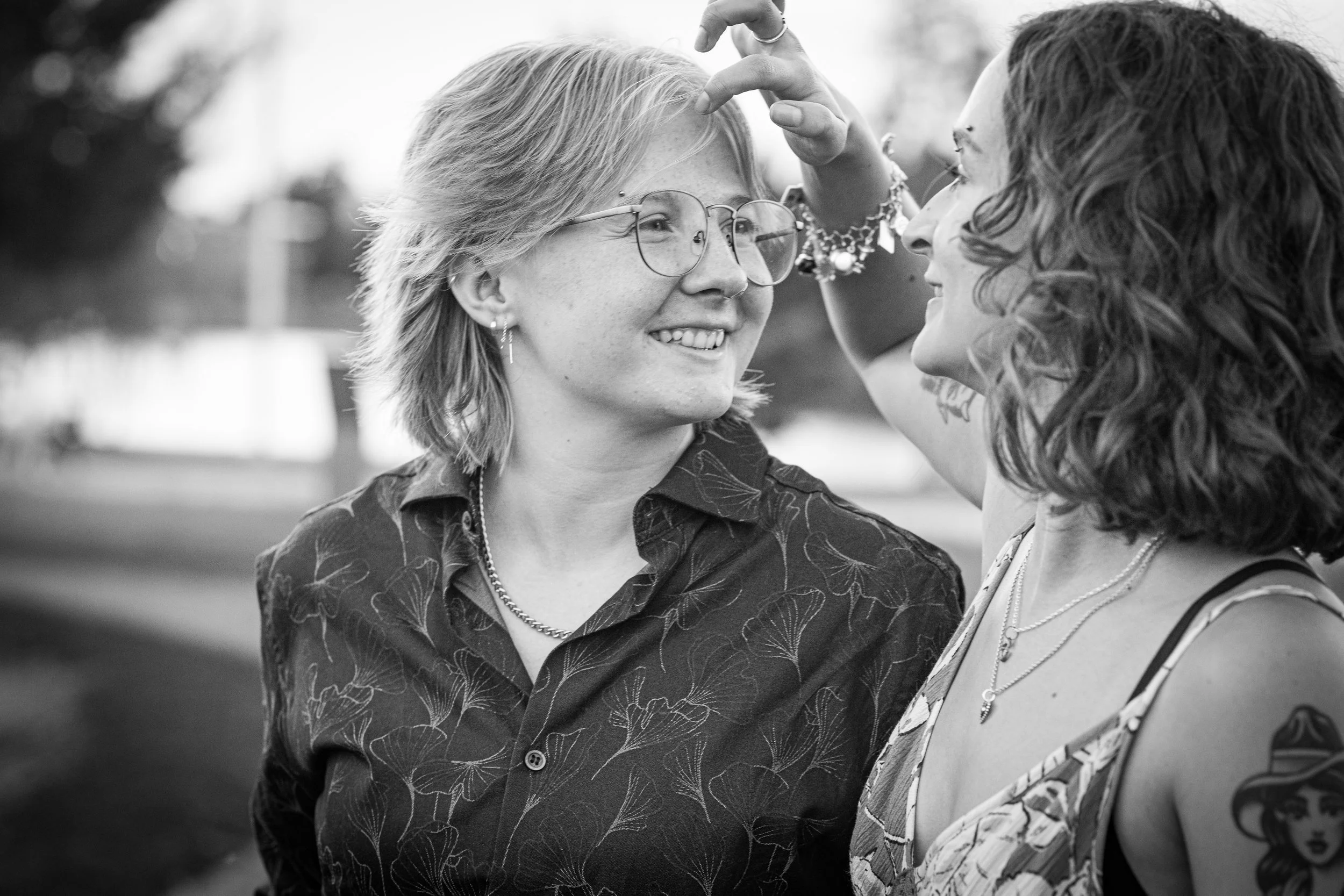 Three women happily interact outdoors, with one touching the other's forehead. The woman in the middle is wearing glasses, earrings, a necklace, and a patterned shirt. The woman on the right has curly hair, layered necklaces, and a tattoo on her arm.