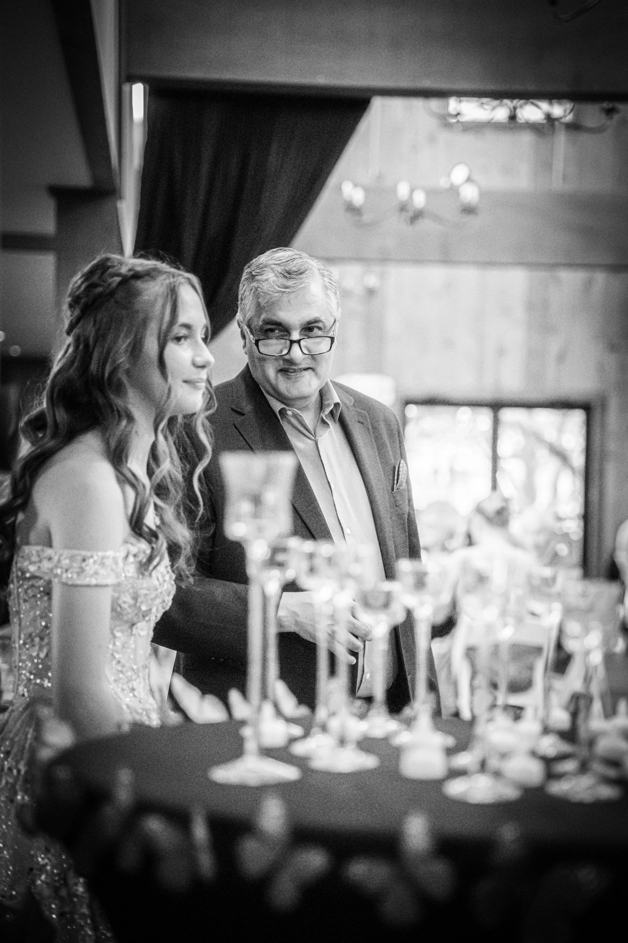 A woman in a sequined dress and an older man in a suit standing by a table with drinks, engaged in conversation at a formal indoor event.