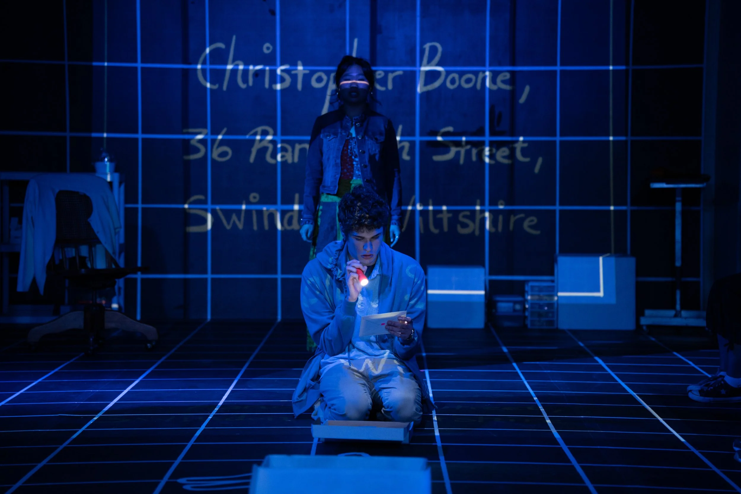 The image depicts a scene in a dark room with blue lighting, featuring two young people. One is sitting on the floor holding a flashlight and reading a piece of paper, while the other stands behind them. The background has a grid pattern on the walls and floor, with handwritten text on the wall indicating an address in England.
