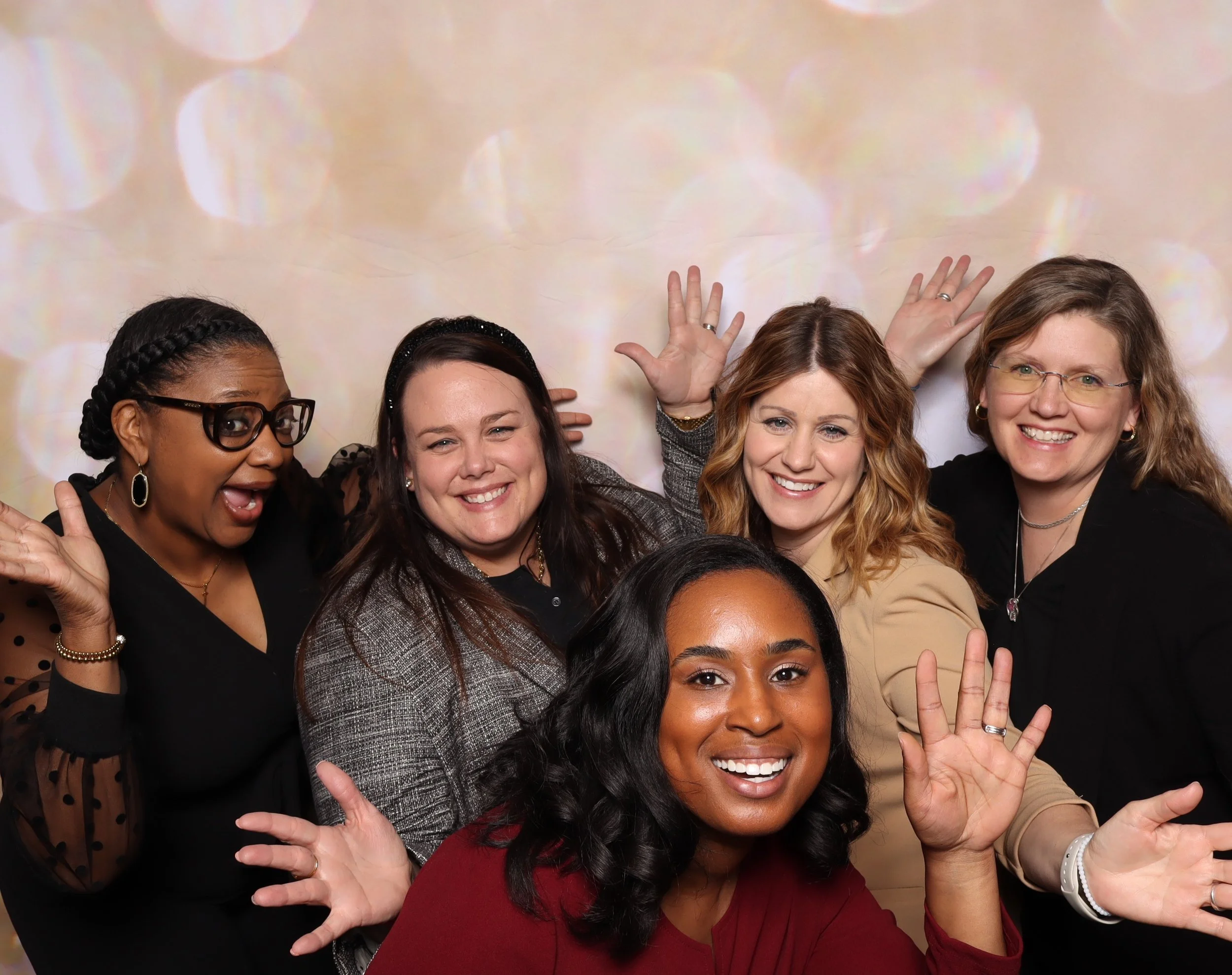 Six women smiling and posing together, waving at the camera, with a colorful bokeh background.