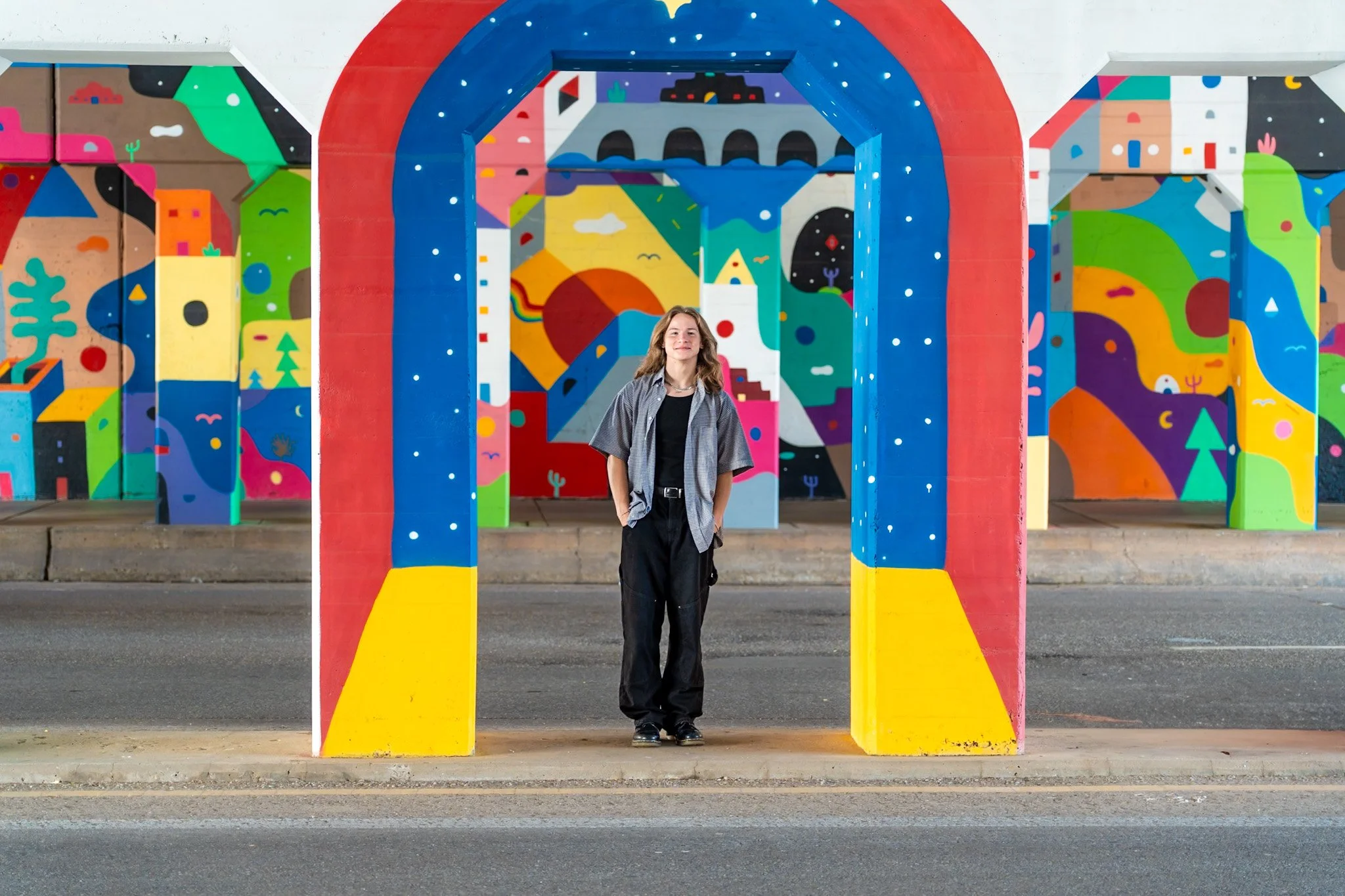 A young woman stands under a colorful, painted urban archway with vibrant, abstract murals on the walls behind her.
