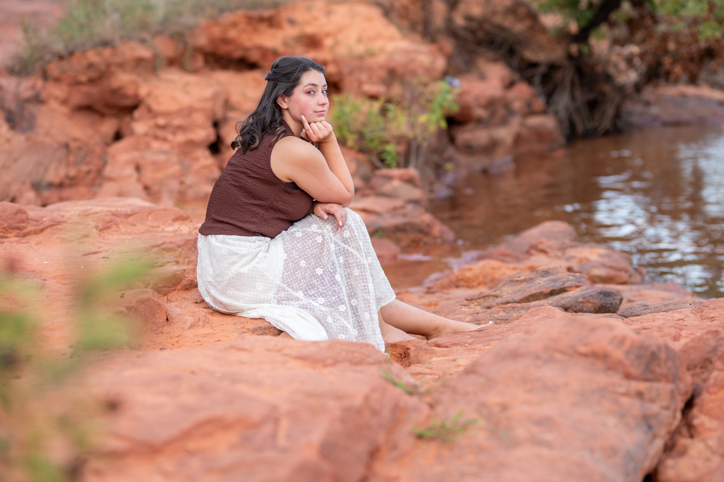 A young woman sitting on red rocks by a body of water, wearing a brown sleeveless top and a white skirt, with her chin resting on her hand and looking thoughtful.