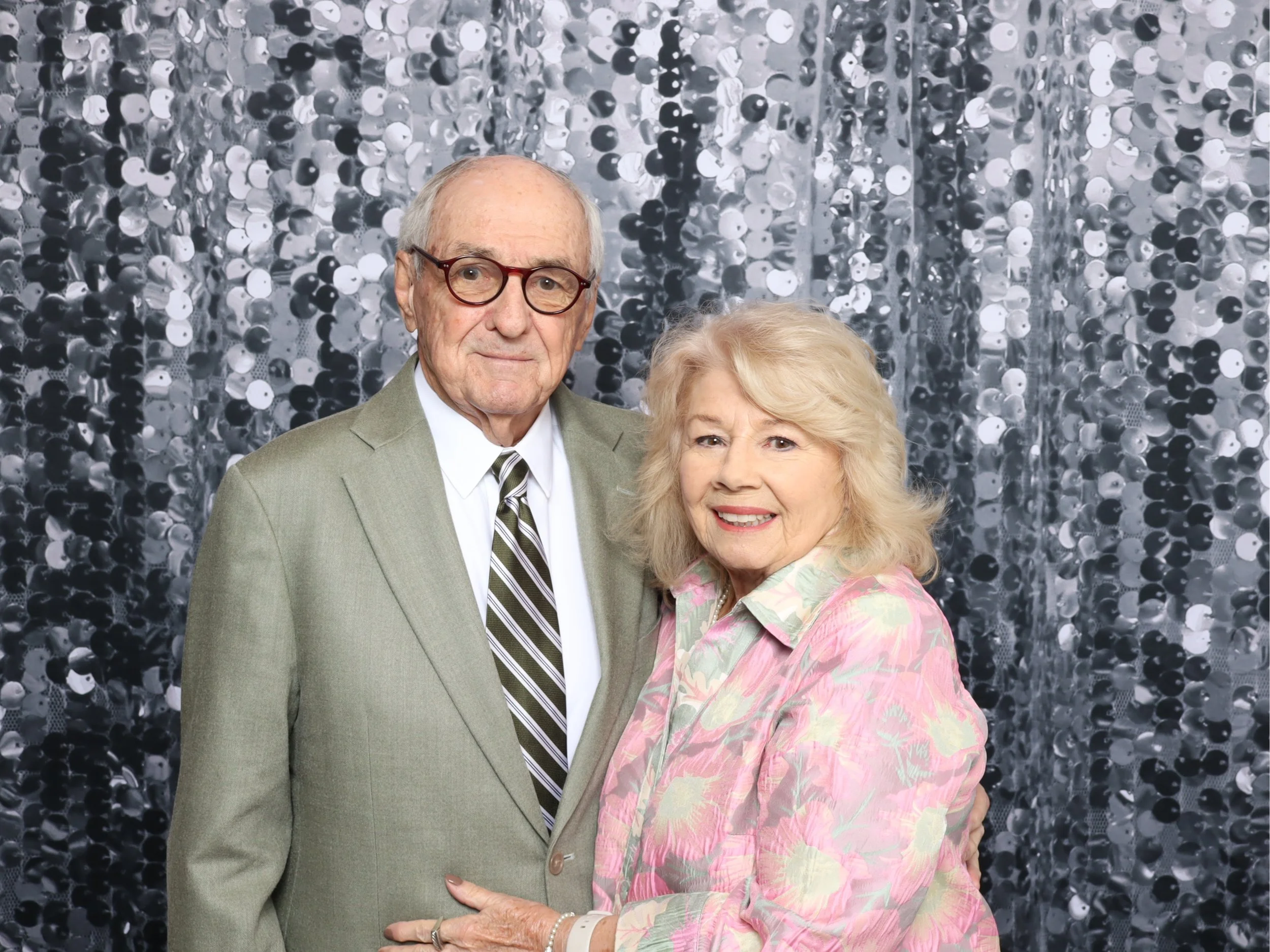 An elderly man and woman standing close together, smiling, in front of a shiny, black and silver sequin backdrop.