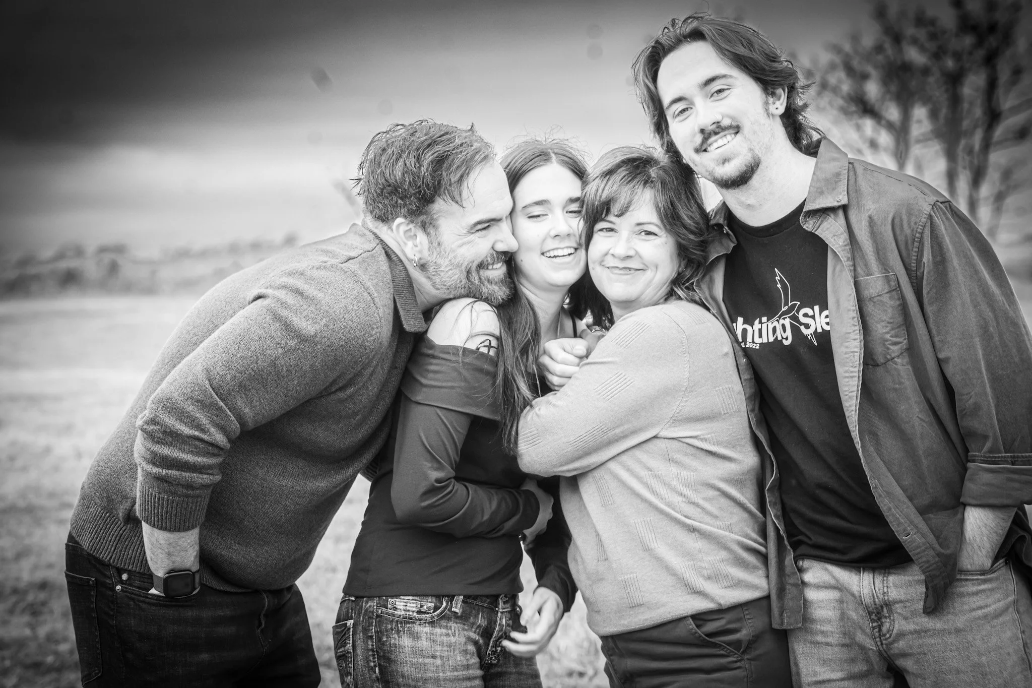 Black and white candid portrait of a family standing close together outdoors, laughing and hugging in a genuine moment of connection