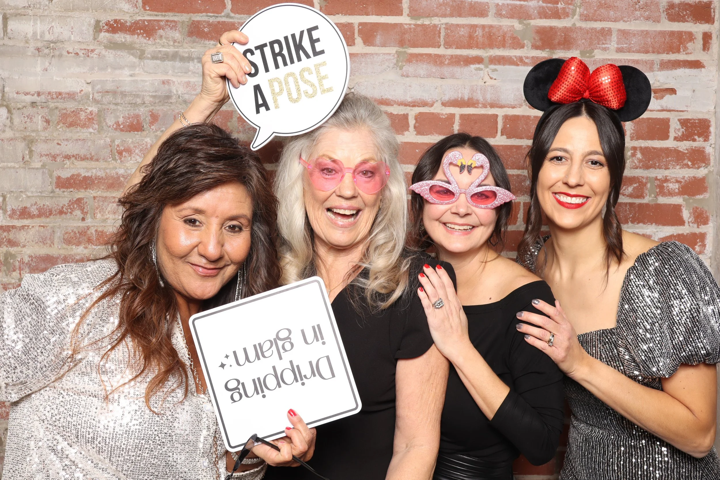 Group of four women celebrating and wearing fun glasses, with two holding signs that read 'Happy in Glam' and 'Strike a Pose', against a brick wall background.
