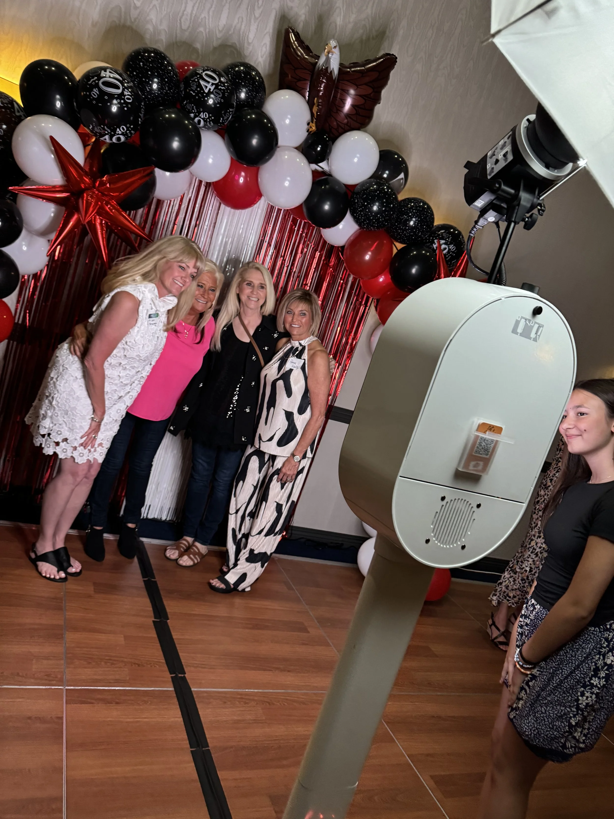 Four women are posing for a photo in front of a backdrop decorated with red, black, and white balloons, including some with the number 40, and a large red star-shaped balloon. A photo booth with a large camera and a young woman taking a picture is visible in the foreground.