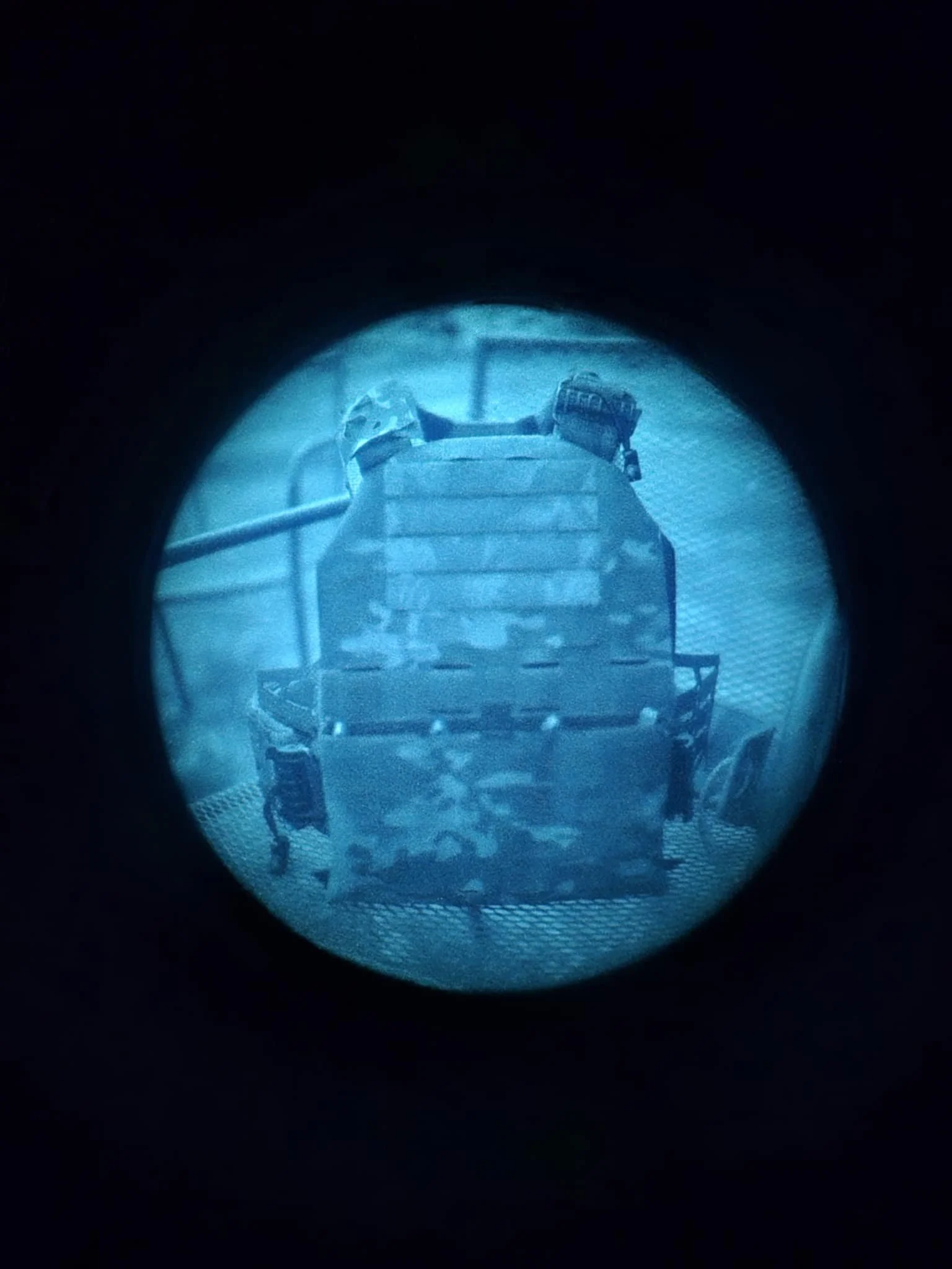 View through a viewfinder showing a military tank with a camouflage pattern, equipped with a mounted weapon, on a metal surface and a railing in the background.
