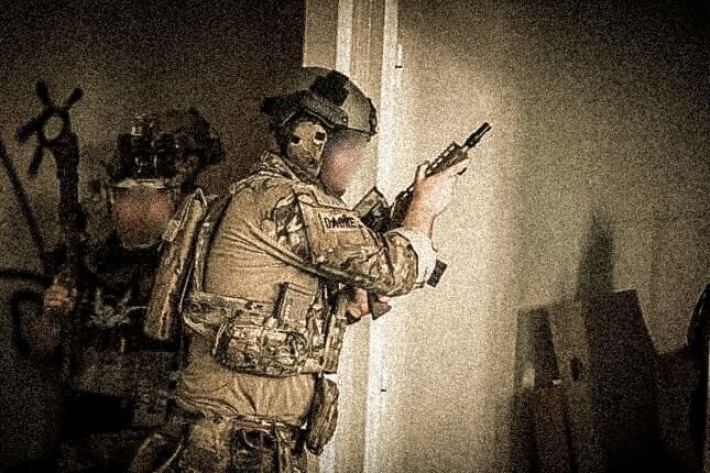A soldier in tan military uniform and helmet aiming a handgun at a wall in an indoor setting.
