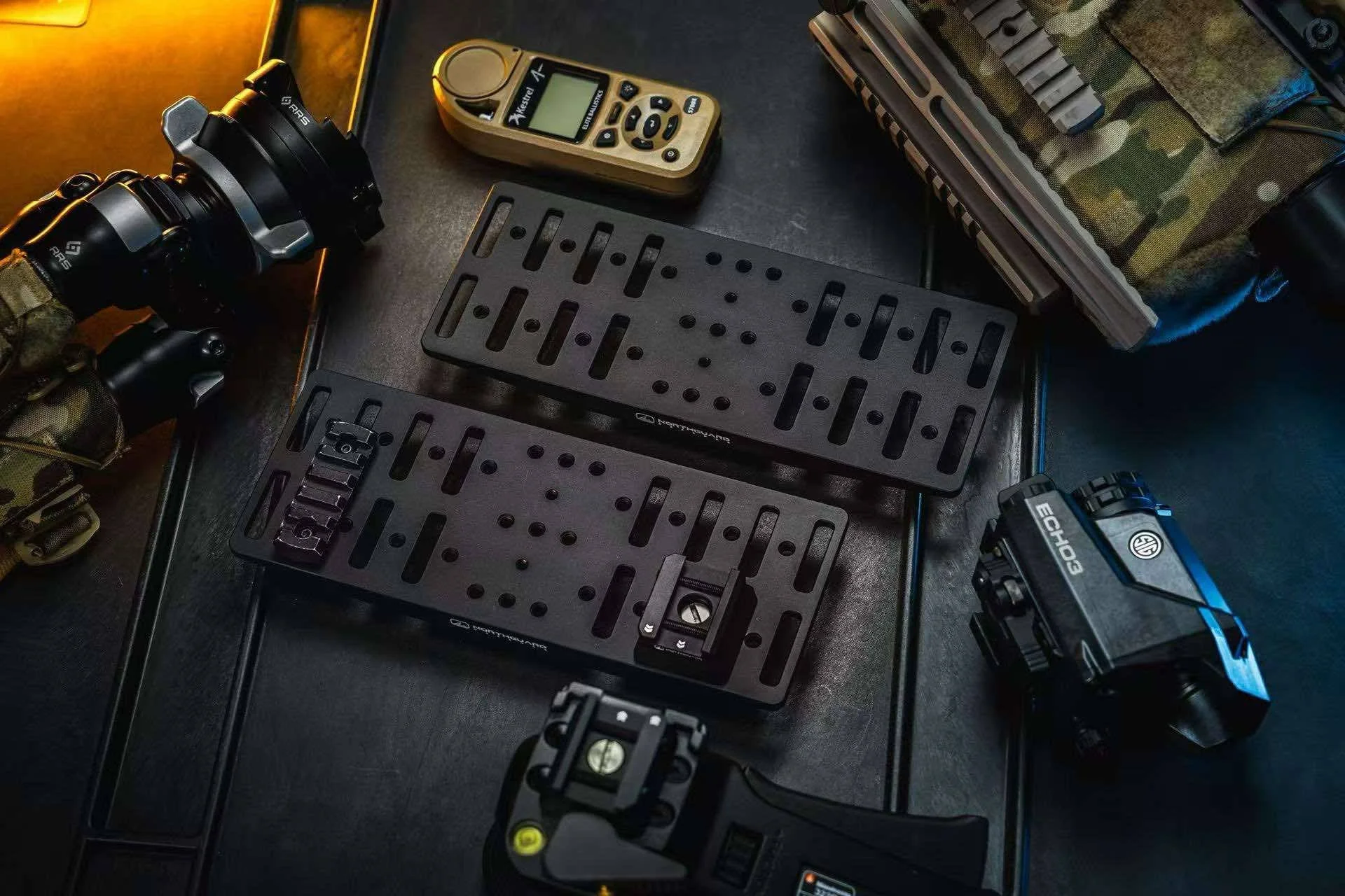 Photography equipment and accessories organized on a dark surface, including a camera, camera's grip, two black camera plates, a GPS device, a handheld laser level, and a camouflaged bag.