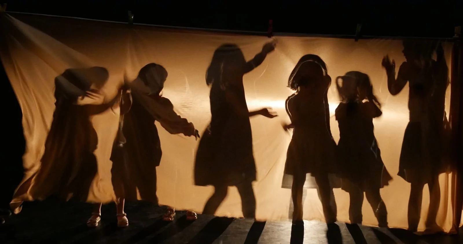 Silhouettes de sept enfants dansant devant un fond lumineux, filmés en ombres chinoises.