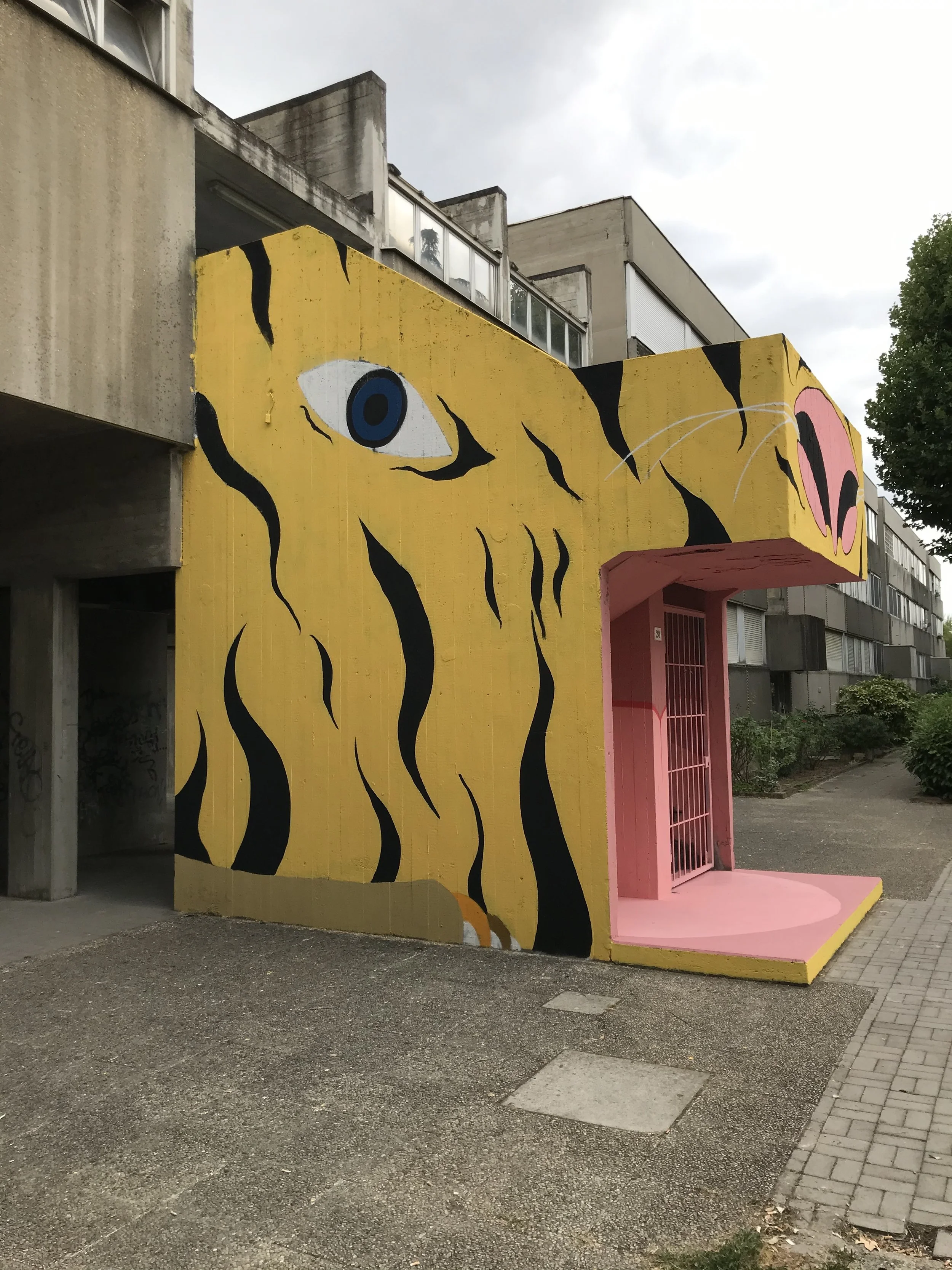 Murale colorata rappresentante una tigre stilizzata con fondo giallo e dettagli neri, con un'area rosa che simula una bocca e alcune linee bianche che rappresentano i baffi.