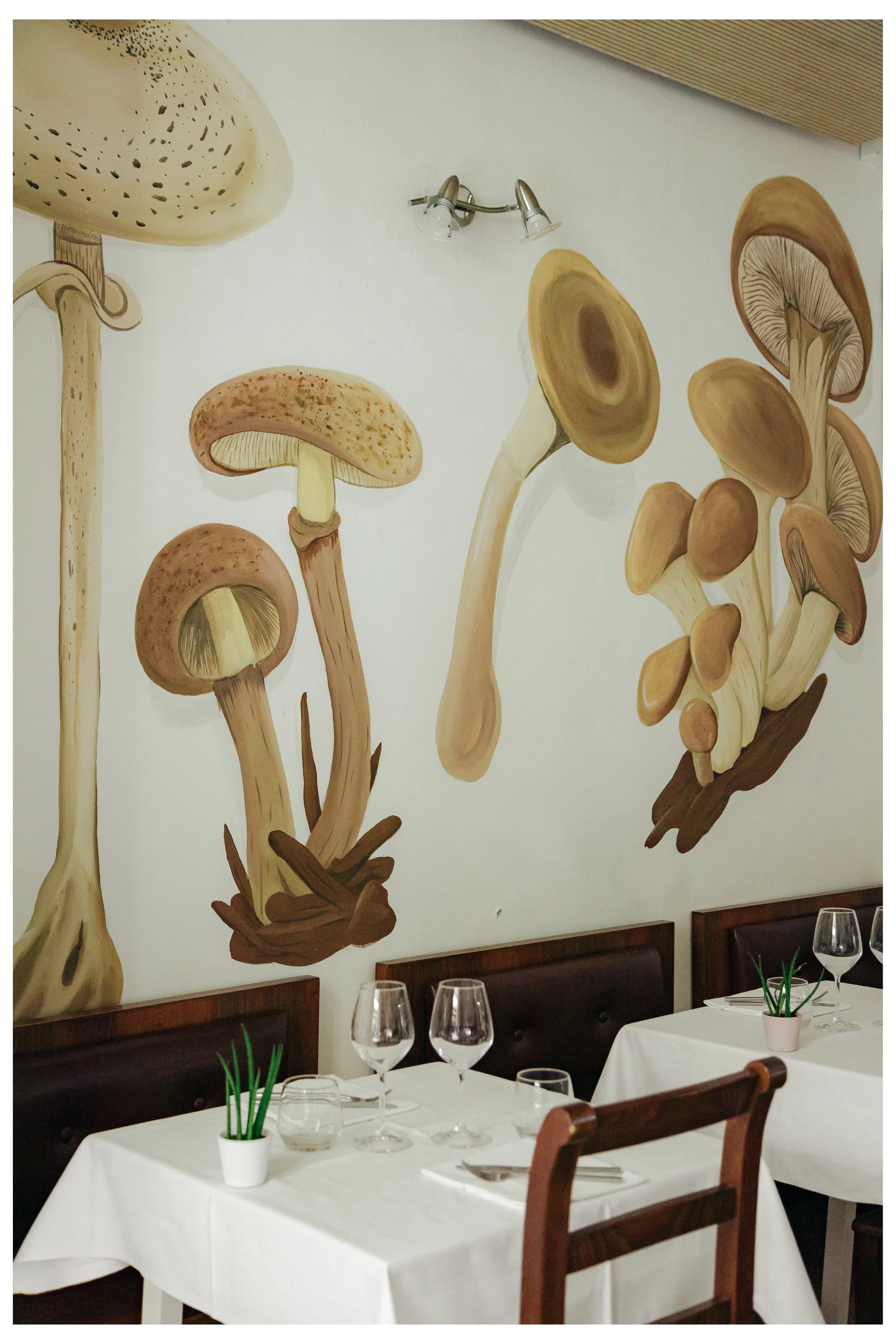 Mural di funghi stilizzati dipinto sulla parete di un ristorante, con tavoli eleganti apparecchiati con tovaglie bianche, bicchieri e posate.