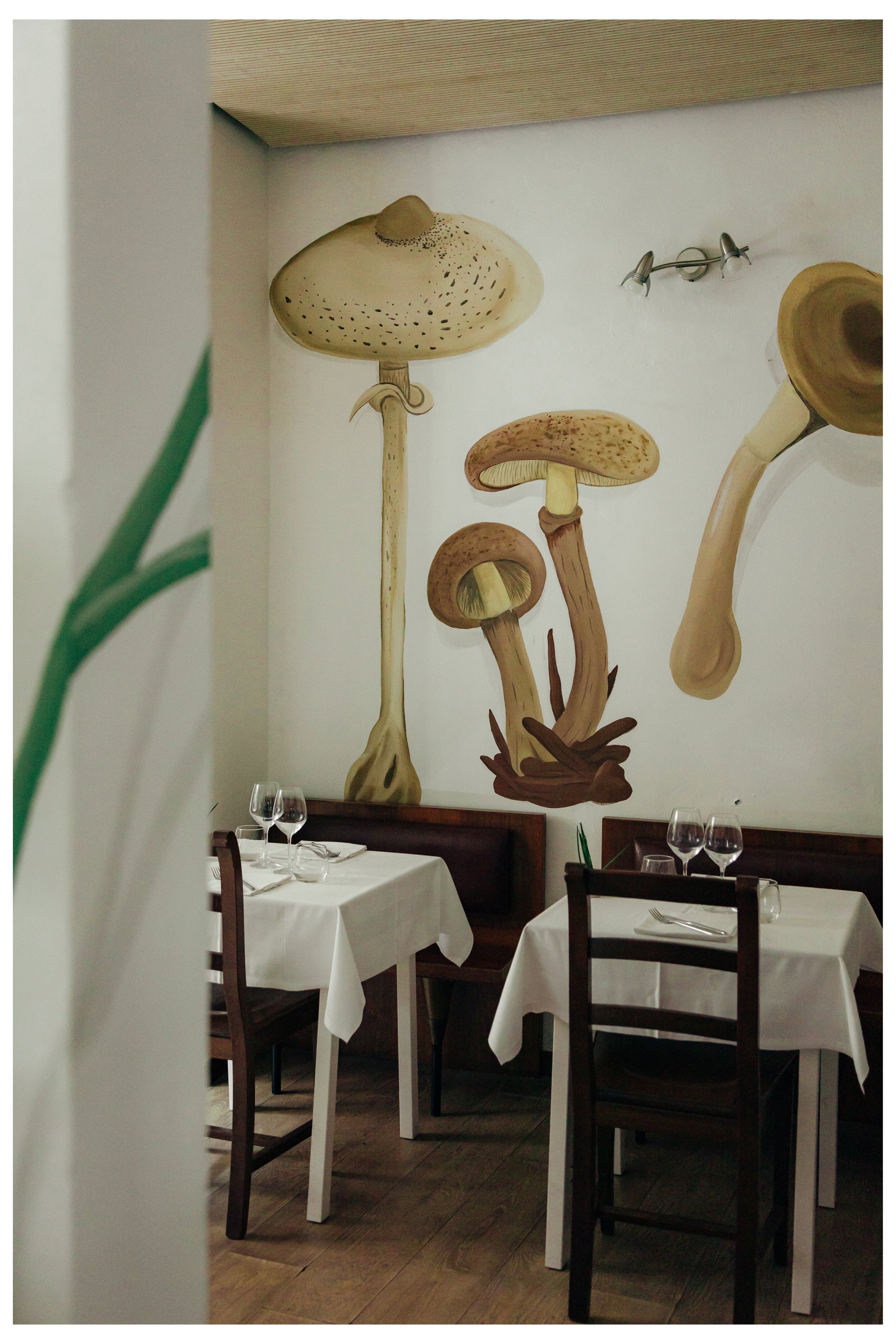 Interno di ristorante con tavoli con tovaglie bianche, bicchieri di vino e posate, parete con decorazione di funghi dipinti