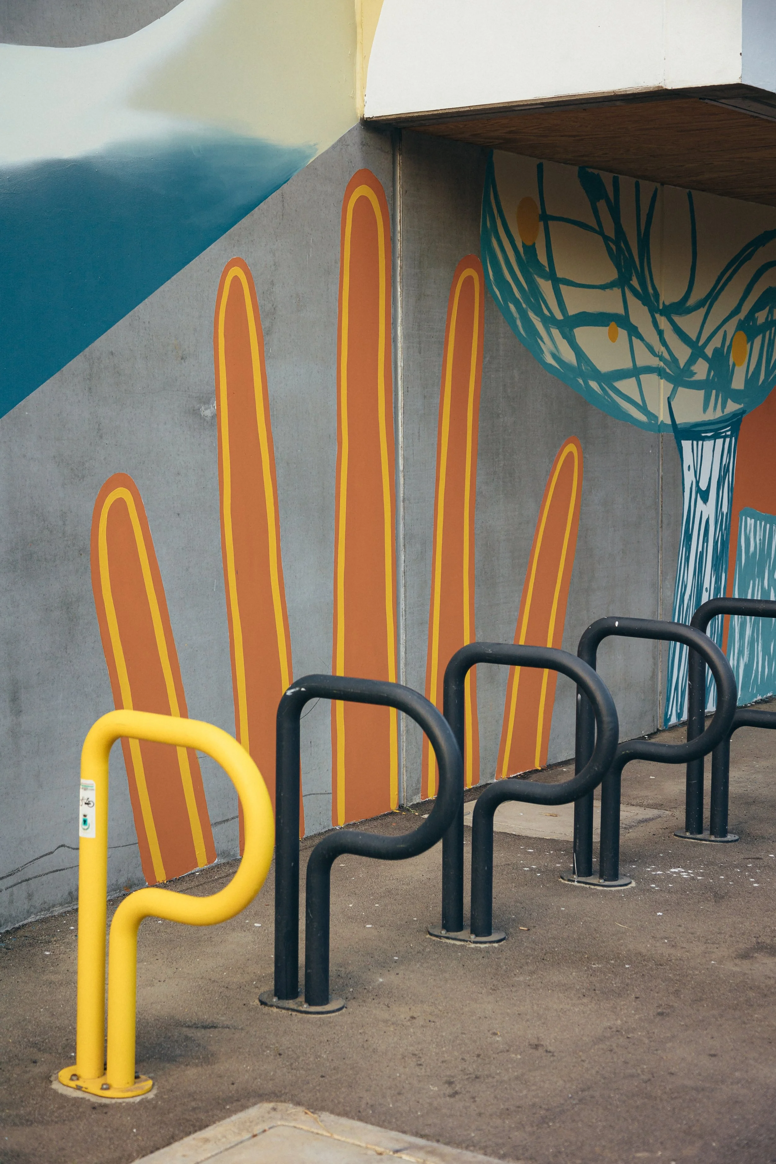 Murale colorata con grandi cactus arancioni e un grande vaso blu con forma di occhio stilizzato, sui muri di un'area urbana, con barriere gialle e nere davanti.