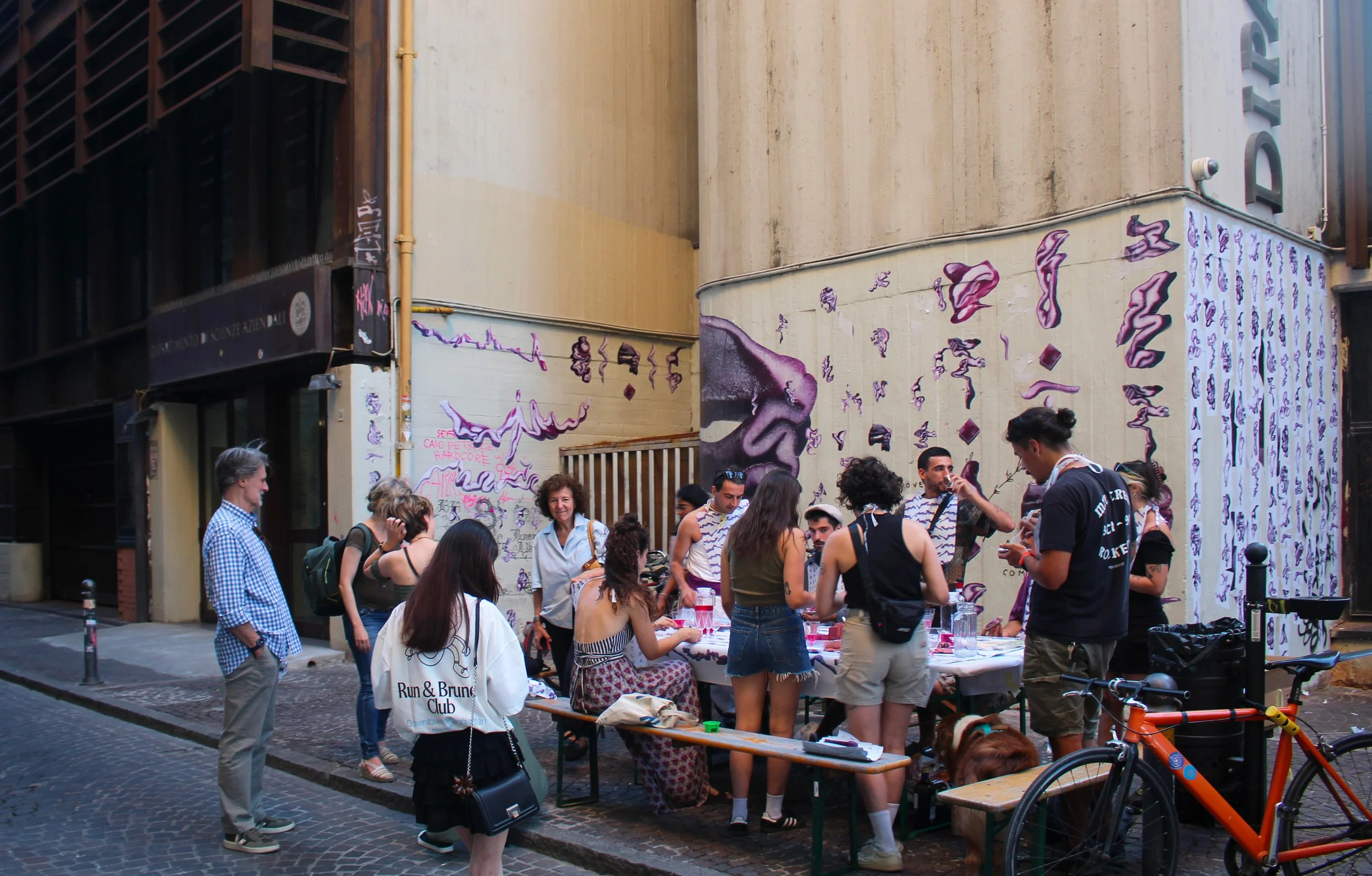 Gruppo di persone che si riuniscono intorno a un tavolo all'aperto in una strada urbana, con murale sulla parete alle spalle e biciclette parcheggiate nei dintorni.