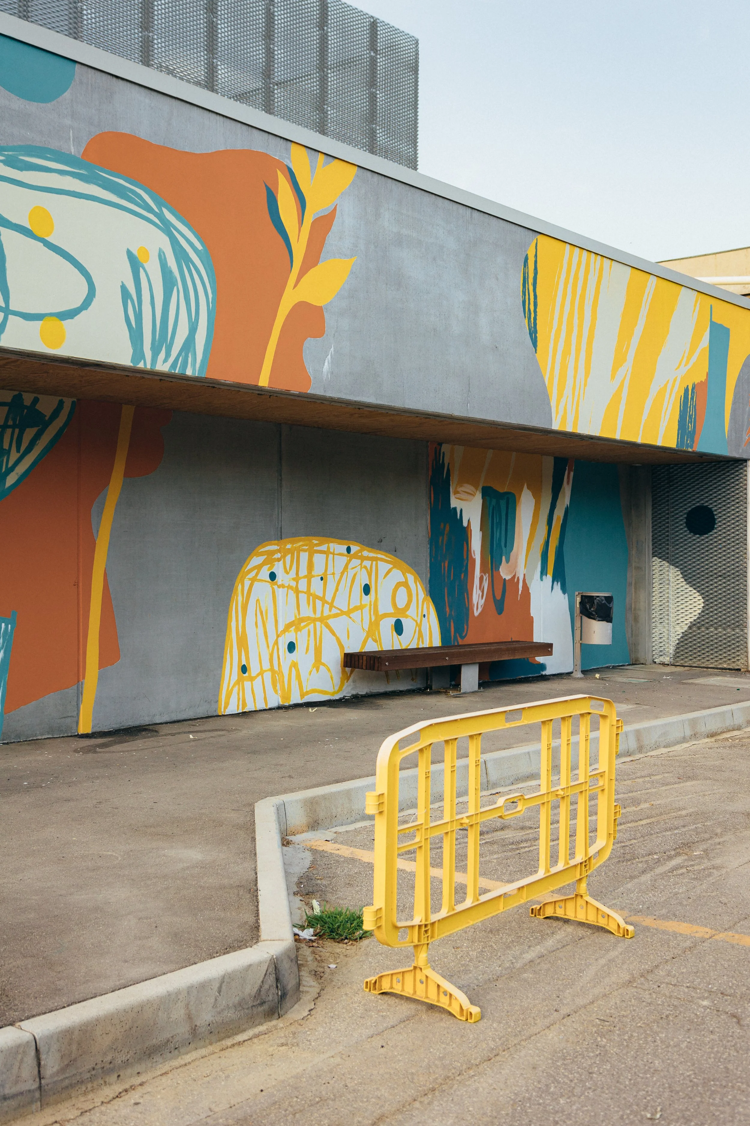 Mural colorato con disegni astratti e elementi di natura, in un'area urbana, con una barriera gialla in primo piano.