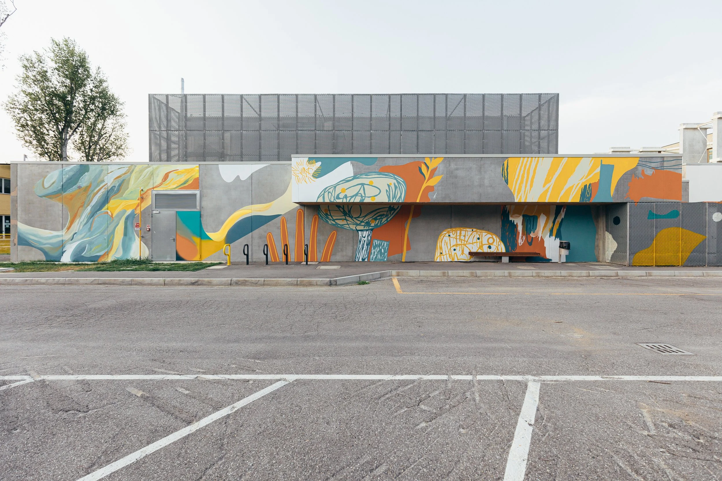 Muro decorato con un murale colorato e astratto, con dettagli di linee e forme organiche, di fronte a una strada e un parcheggio.