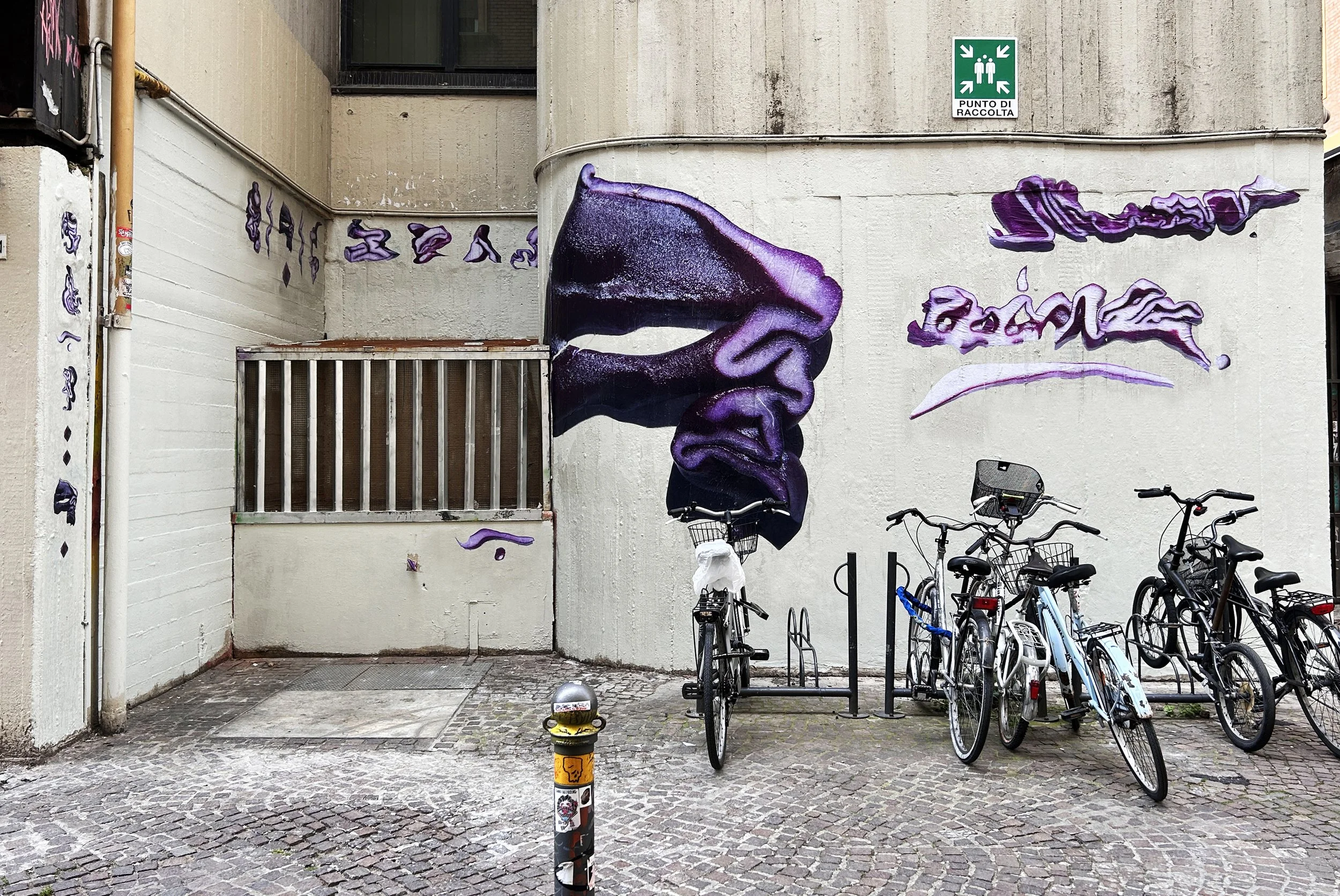 Murale che raffigura un grande bicchiere di vino rosso con sfumature violacee, adornerato con dettagli artistici viola e nero, e testo stilizzato. In alto a destra, segnale di raccolta punti sulla parete.