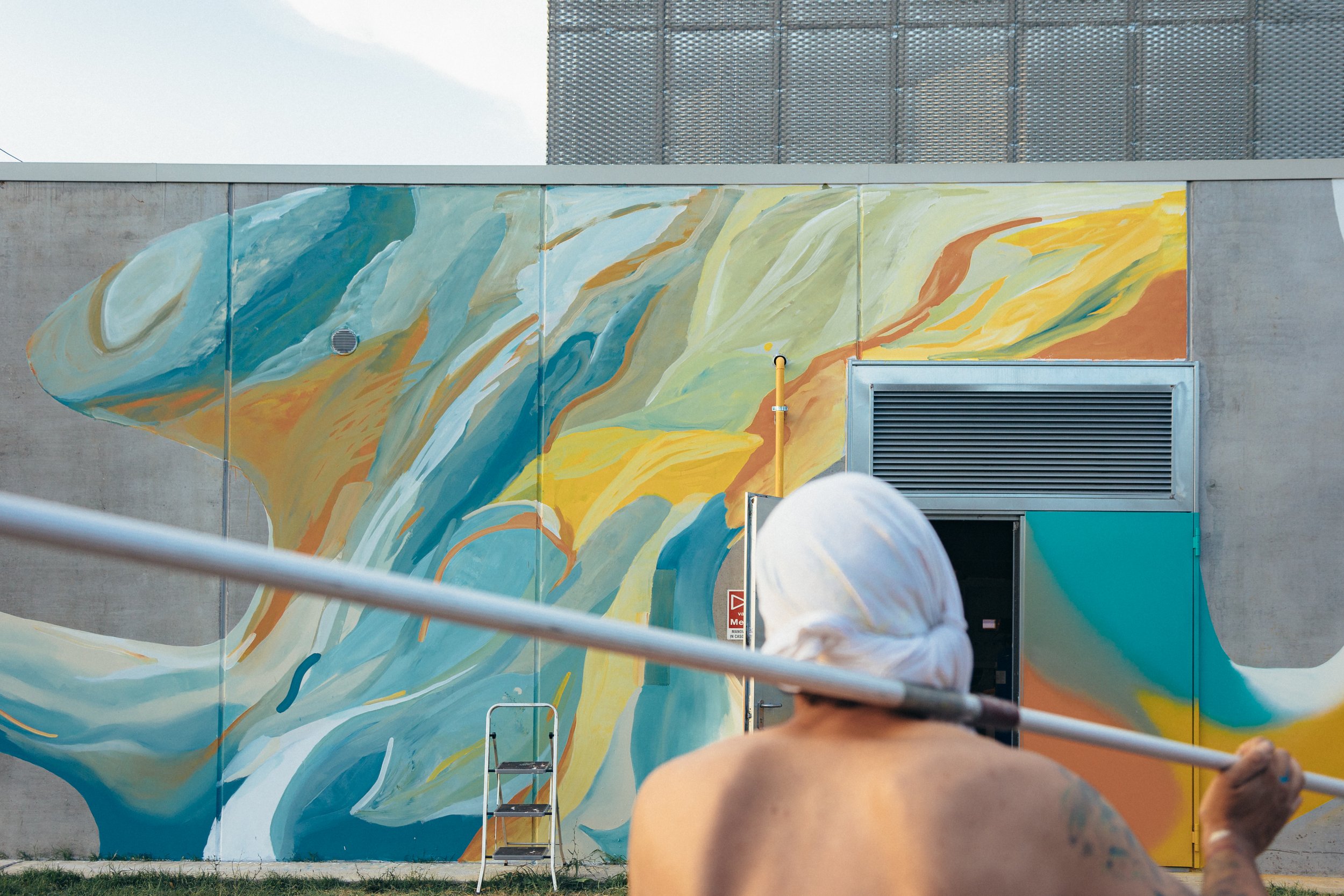 Un mur con uno dipinto astratto di colori blu, verde, giallo e marrone, con alcune persone che passato davanti, una delle quali tiene in mano un pennello.
